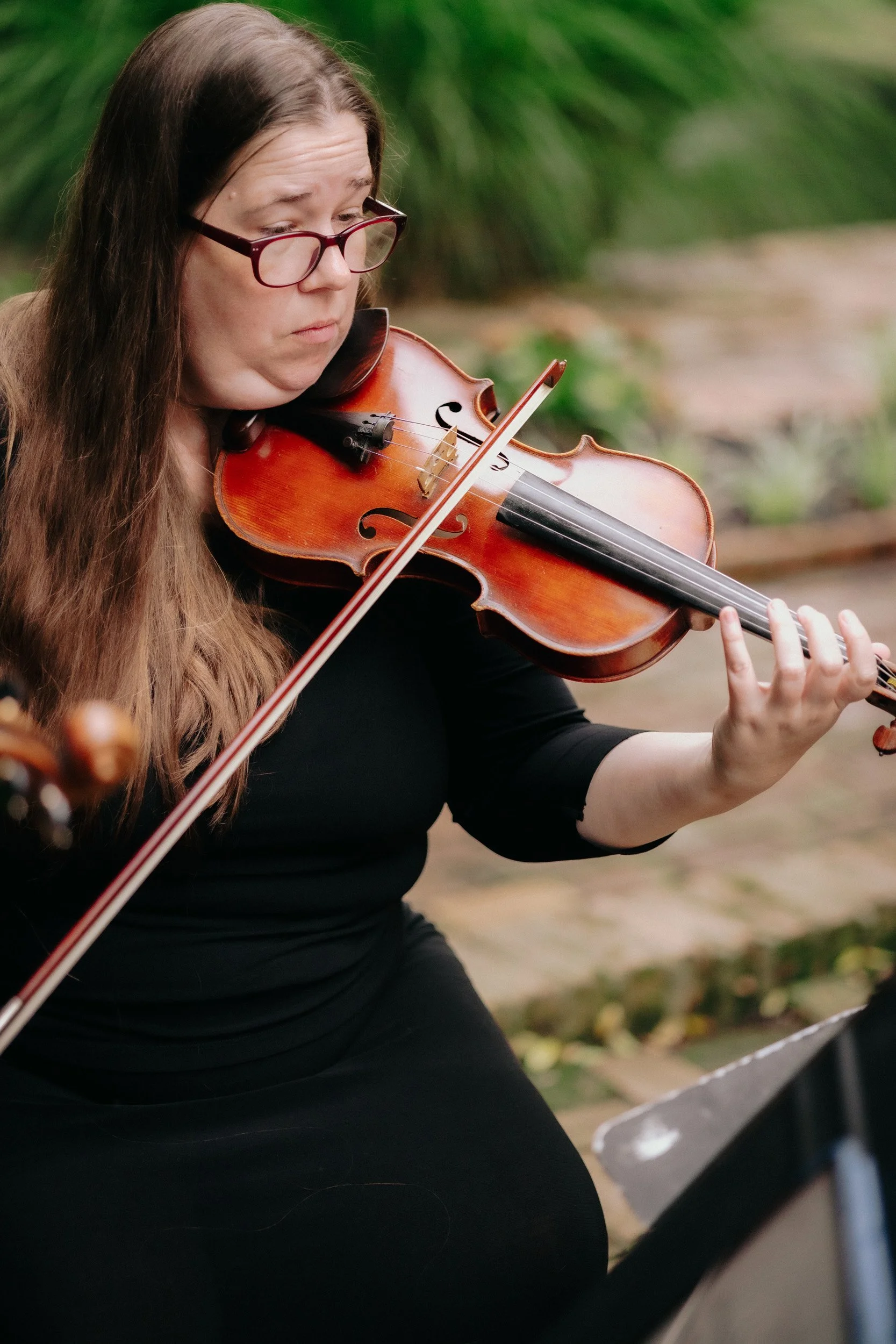 A woman with long brown hair, wearing glasses, playing a violin outdoors.