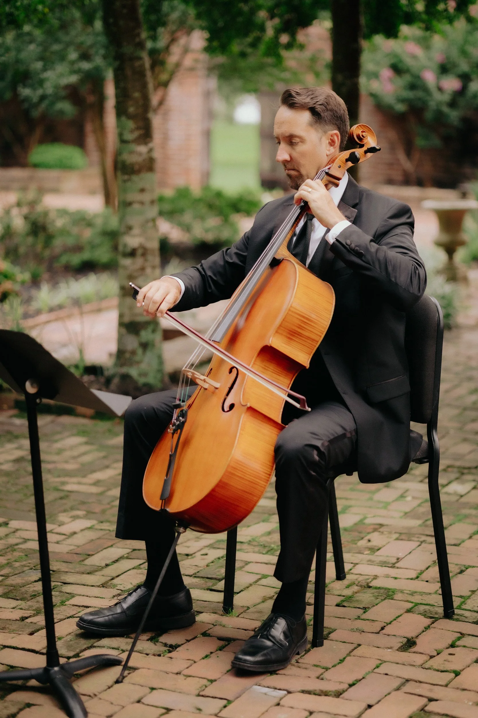 A man in a formal suit playing a cello outdoors in a garden or park setting.
