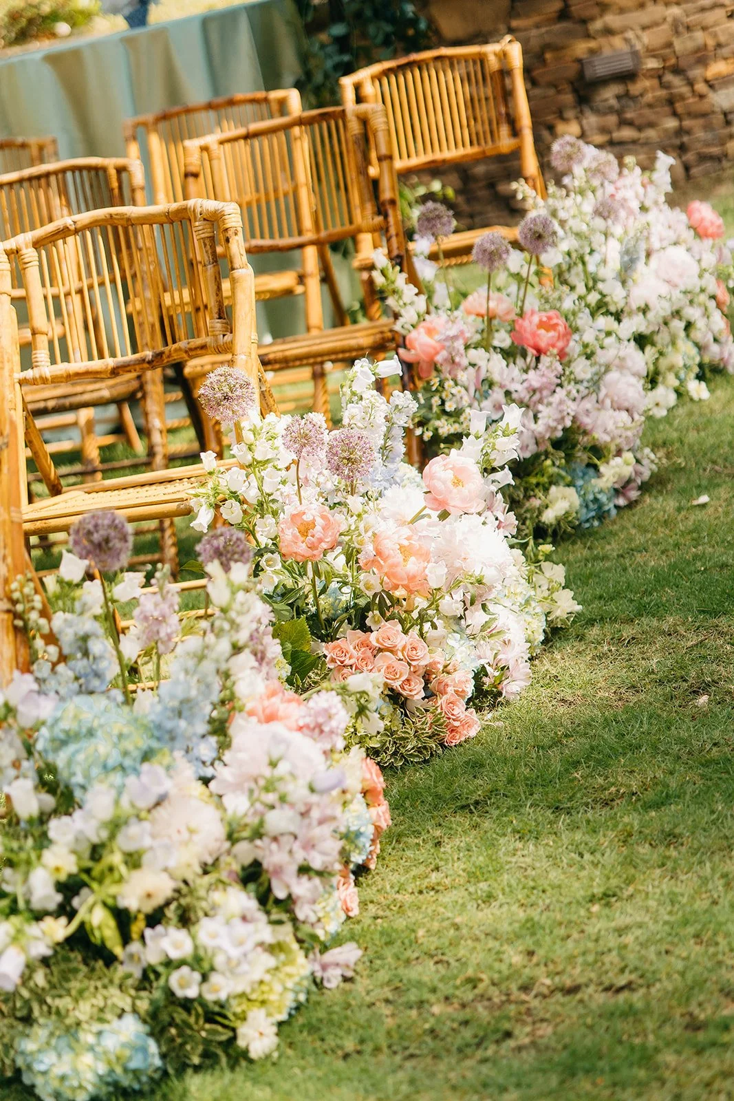 Wooden chairs lined up with a floral arrangement in front, including pink, white, and purple flowers, on a grassy lawn at an outdoor event.