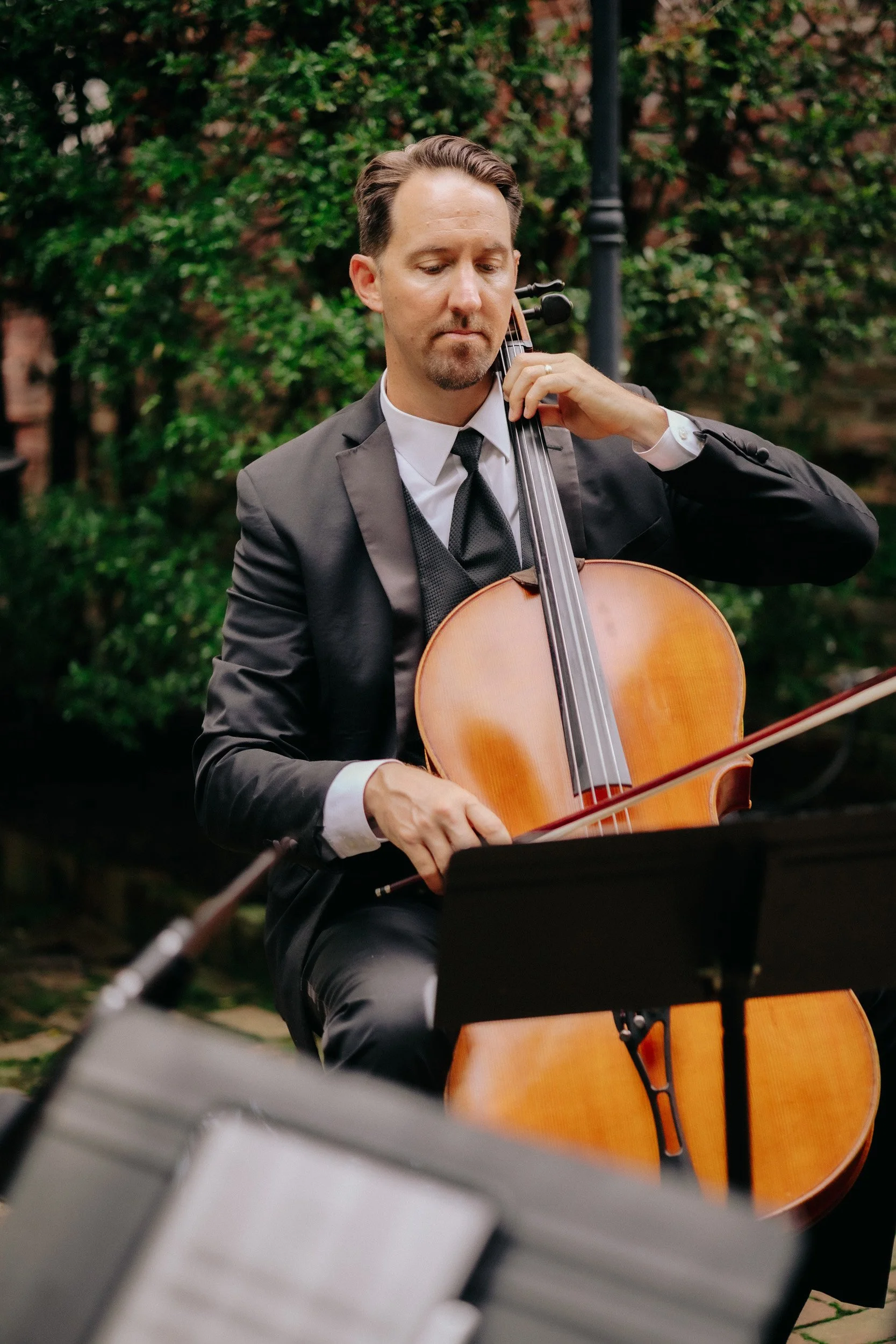 A man in a black suit, white shirt, and black tie playing a cello outdoors, with greenery and a lamp post in the background.