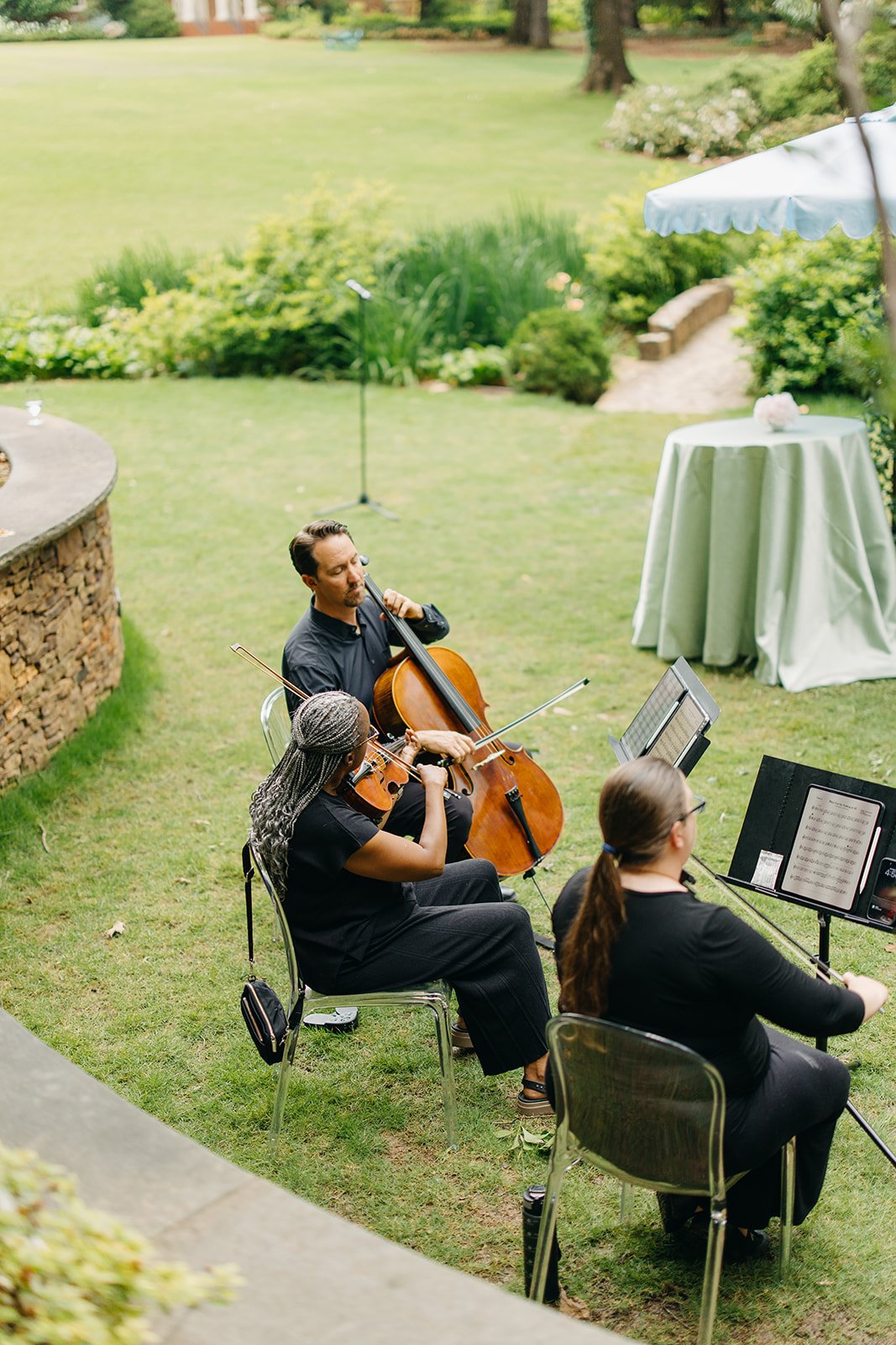 Three musicians playing string instruments outdoors in a garden, with a cello, violin, and viola, near a table with a white tablecloth and an umbrella, surrounded by greenery.