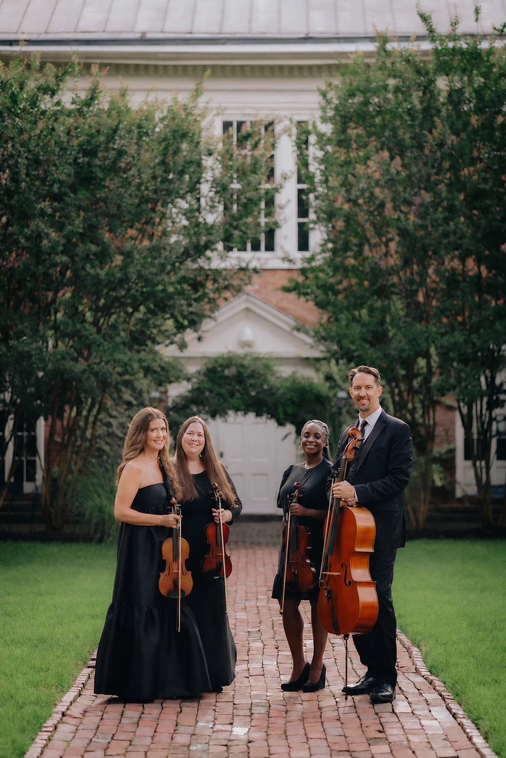 A group of five musicians in formal black attire holding string instruments, standing on a brick pathway in front of a house with green trees in the background.