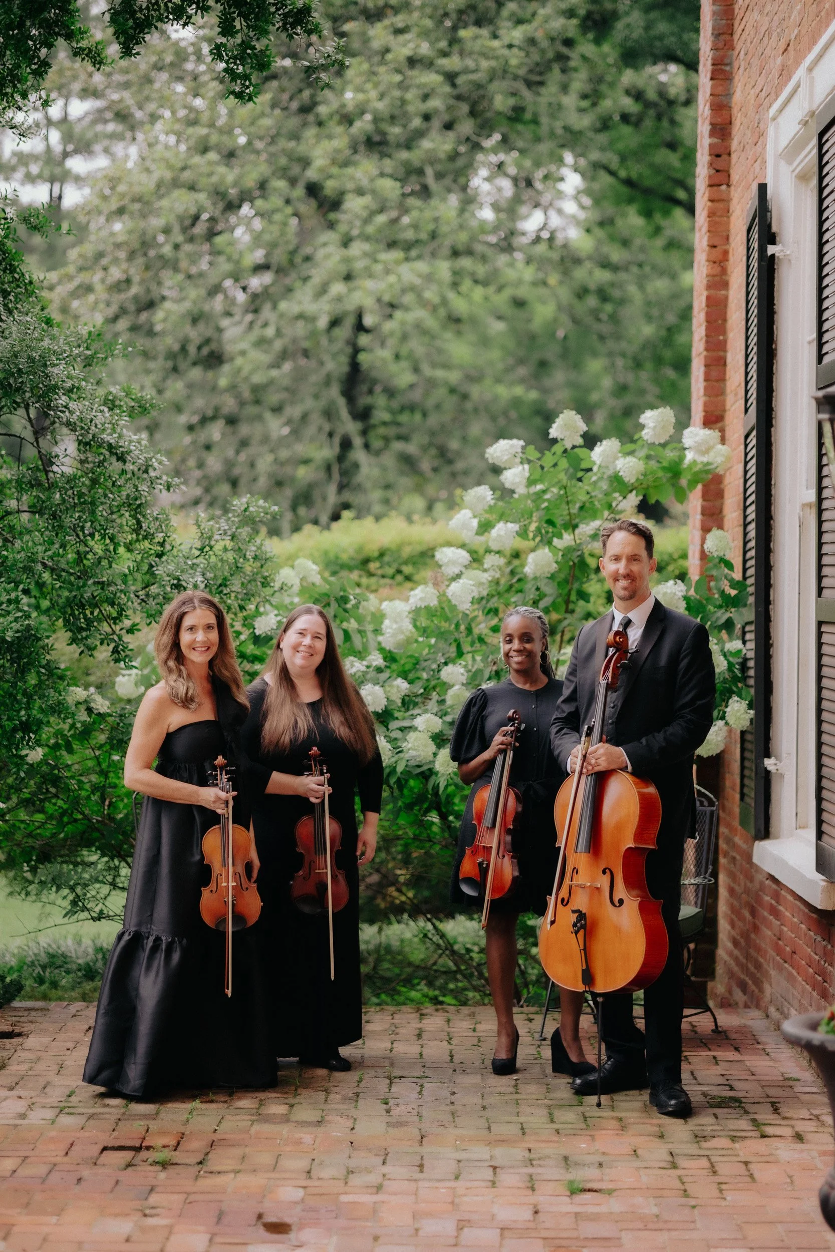 A group of five musicians holding string instruments, three women and two men, standing outdoors on a brick patio surrounded by green foliage and blooming white flowers, with a brick building on the right side.