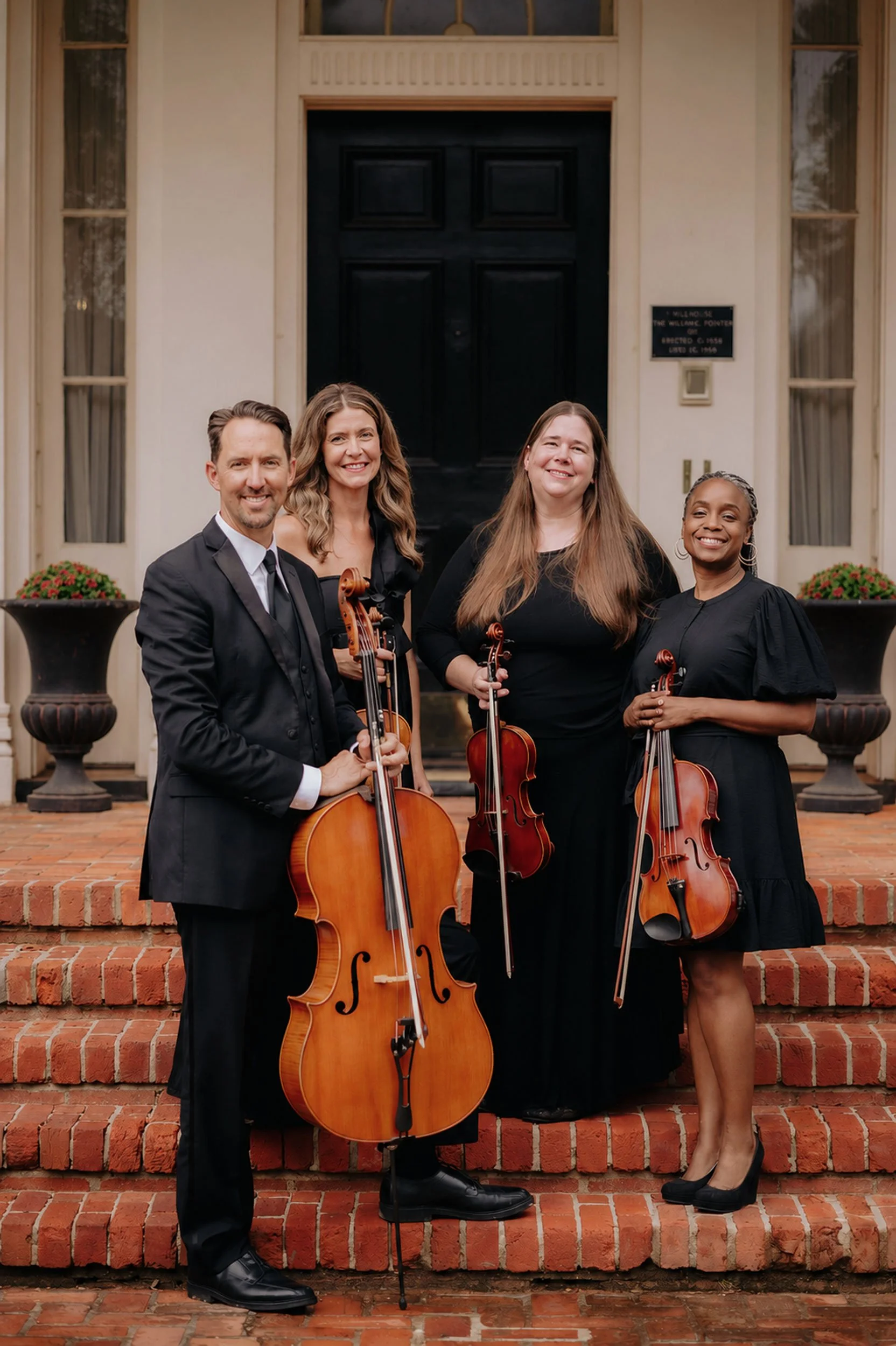 Group of four people holding violins and posing on brick steps in front of a building with black door and plaques.