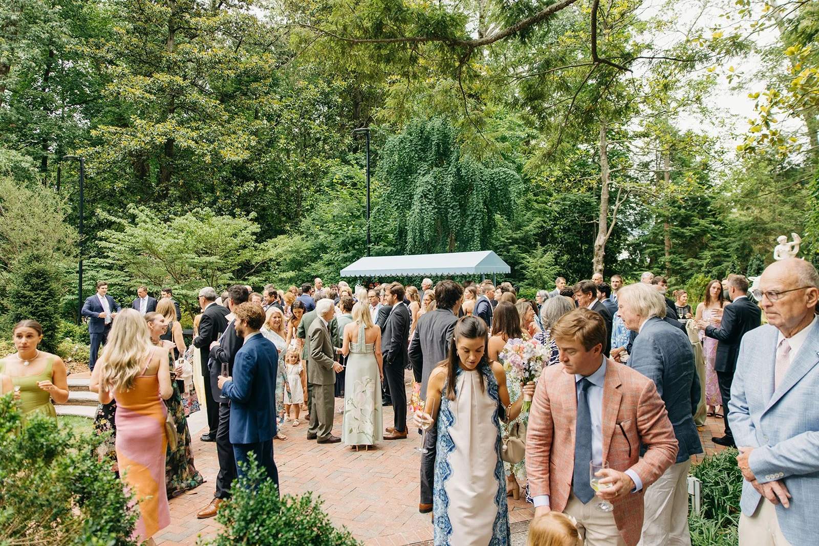 A large group of people dressed formally at an outdoor garden event, with trees and greenery in the background.