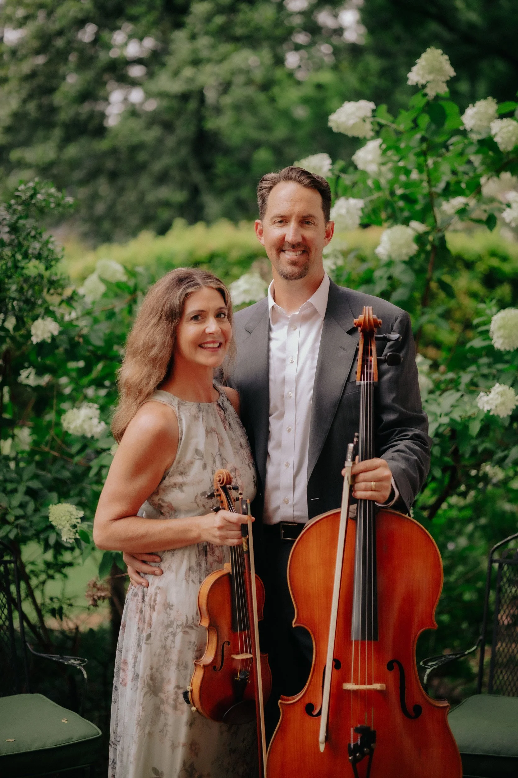A man and woman standing outdoors surrounded by greenery, holding string instruments, with the man in a suit and the woman in a floral dress.