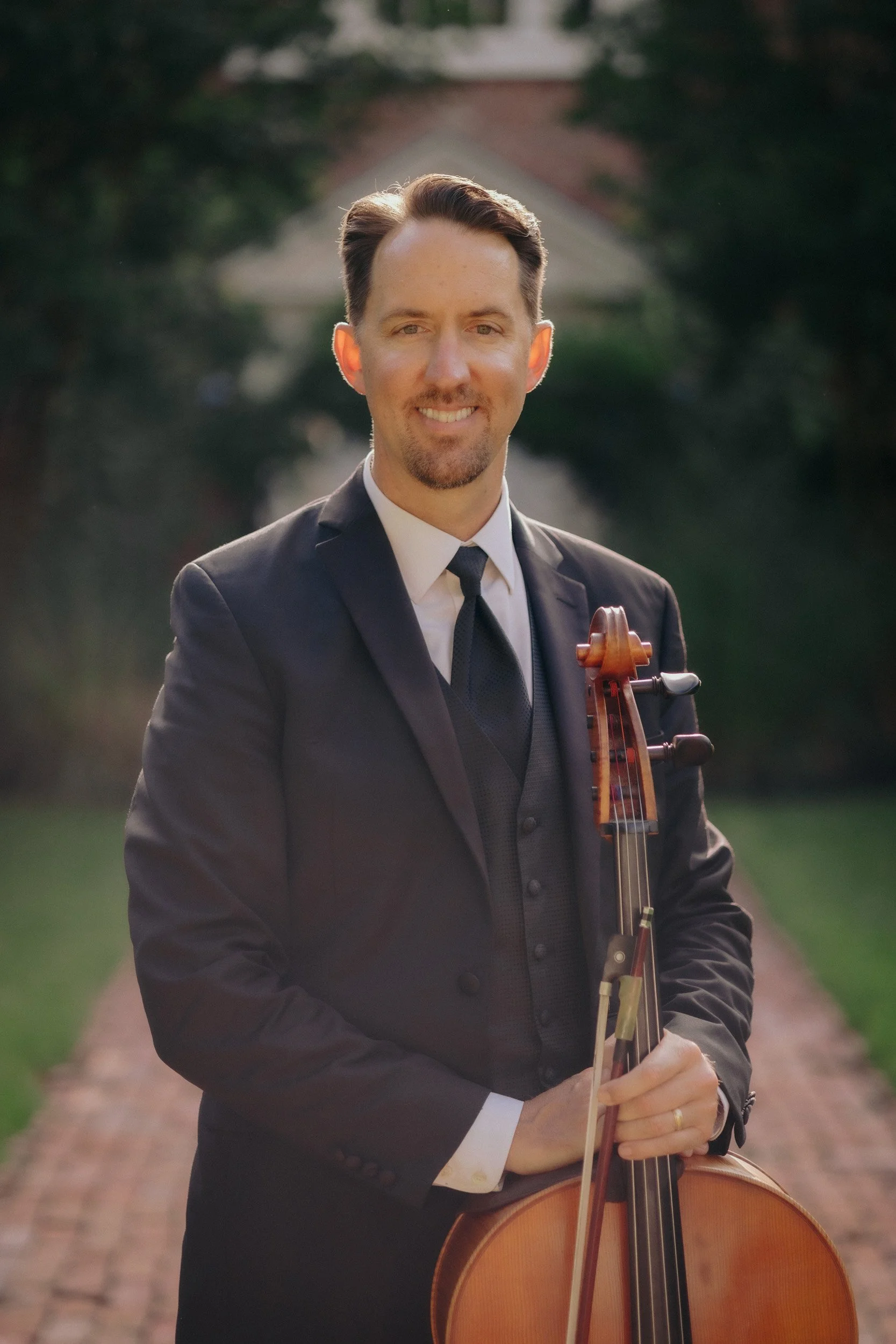 A man in a suit holding a cello outdoors on a path with trees in the background.
