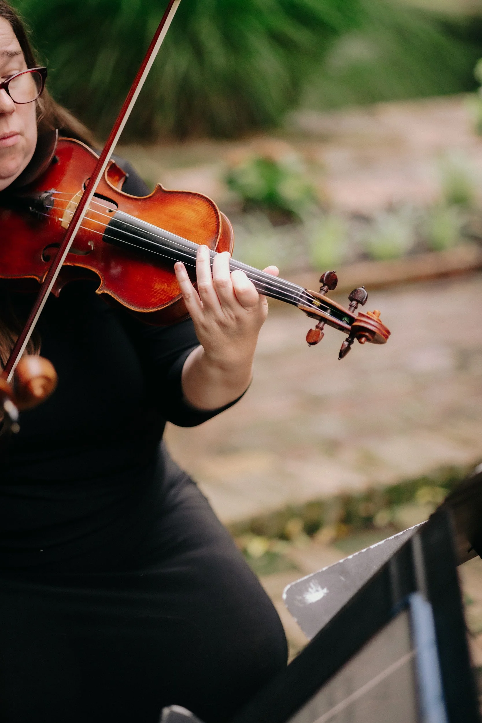 A person playing the violin outdoors, seated on a bench, surrounded by greenery.
