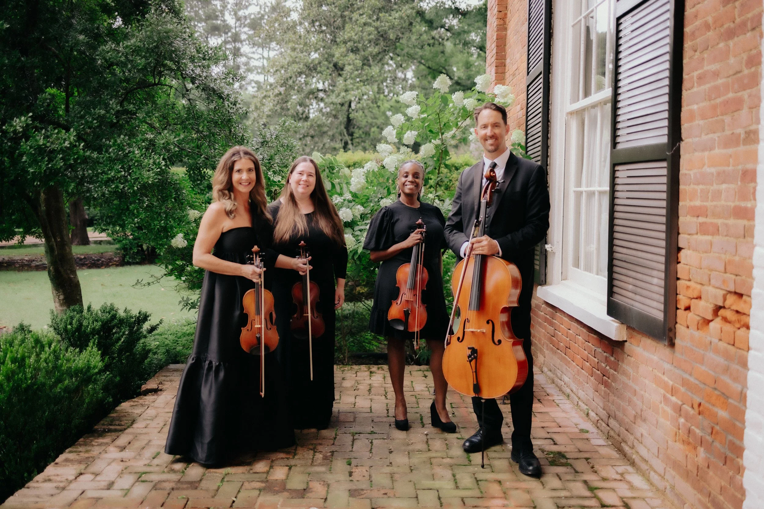 Four musicians posing outdoors with string instruments, standing on a brick pathway beside a brick building with black shutters, surrounded by greenery and flowering bushes.