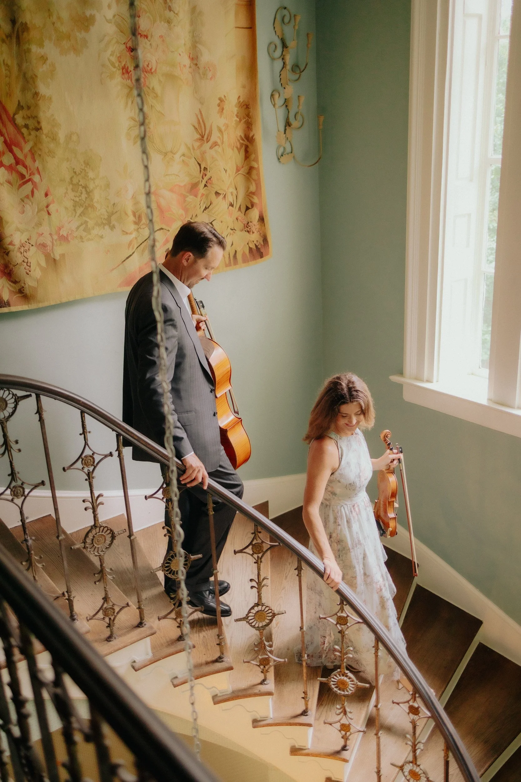A man in a suit and a woman in a floral dress standing on a staircase, both holding violins, with the man looking down and the woman smiling.