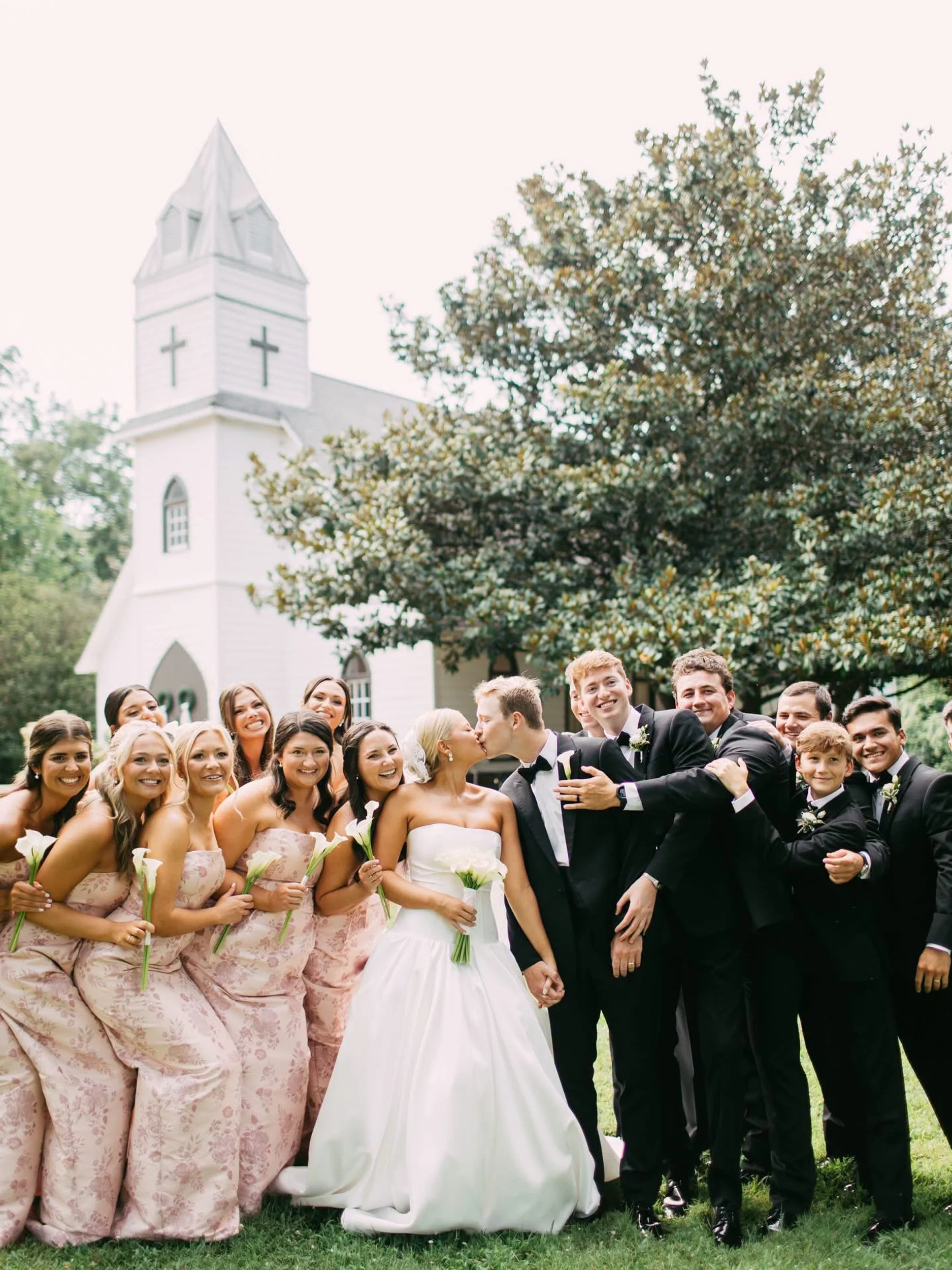 A wedding celebration with a bride and groom kissing, surrounded by wedding party members including bridesmaids in pink floral dresses and groomsmen in black tuxedos, outdoors in front of a white church with a steeple and trees.