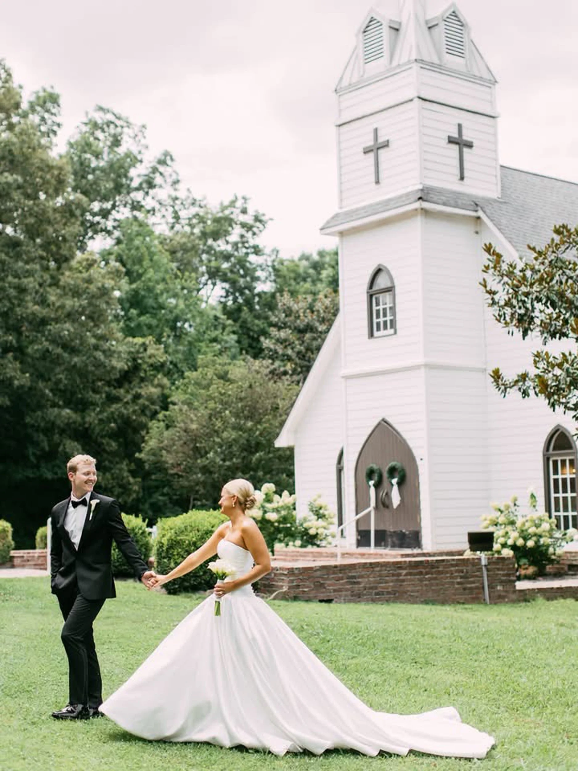 A newlywed couple holding hands and smiling in front of a white church with a steeple, decorated with wreaths, during daytime. The bride is in a strapless wedding gown with a long train, and the groom in a black tuxedo.