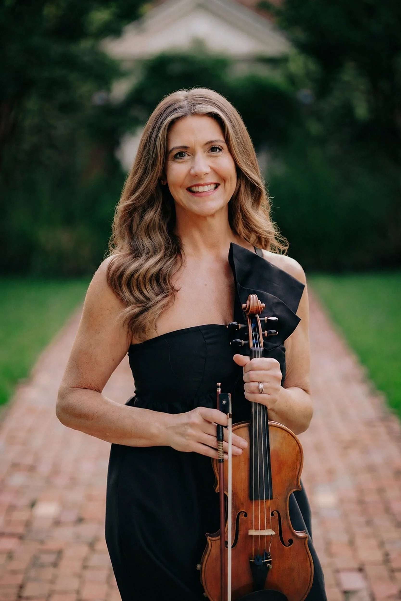 A woman with long, wavy brown hair smiling while holding a violin and bow outdoors on a brick pathway, with greenery and trees in the background.