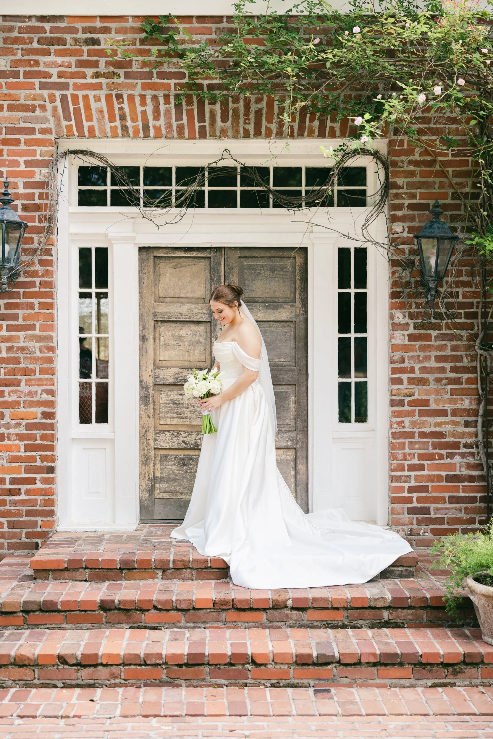 A bride in a white wedding gown holding a bouquet of white flowers standing on brick stairs in front of a wooden door and brick wall, with greenery and pink flowers overhead.