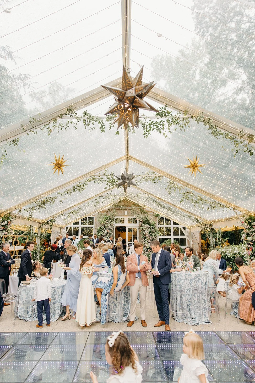 A wedding reception inside a decorated glass-roofed venue with hanging star-shaped lights and greenery, guests gathered around tables, and children in the foreground.