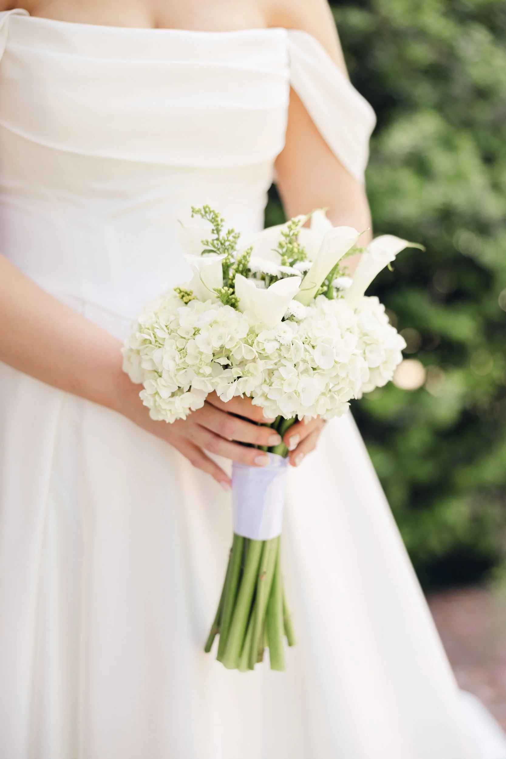 Close-up of a bride in a white wedding dress holding a bouquet of white flowers, including calla lilies and roses, with green foliage, outdoors with blurred greenery in the background.
