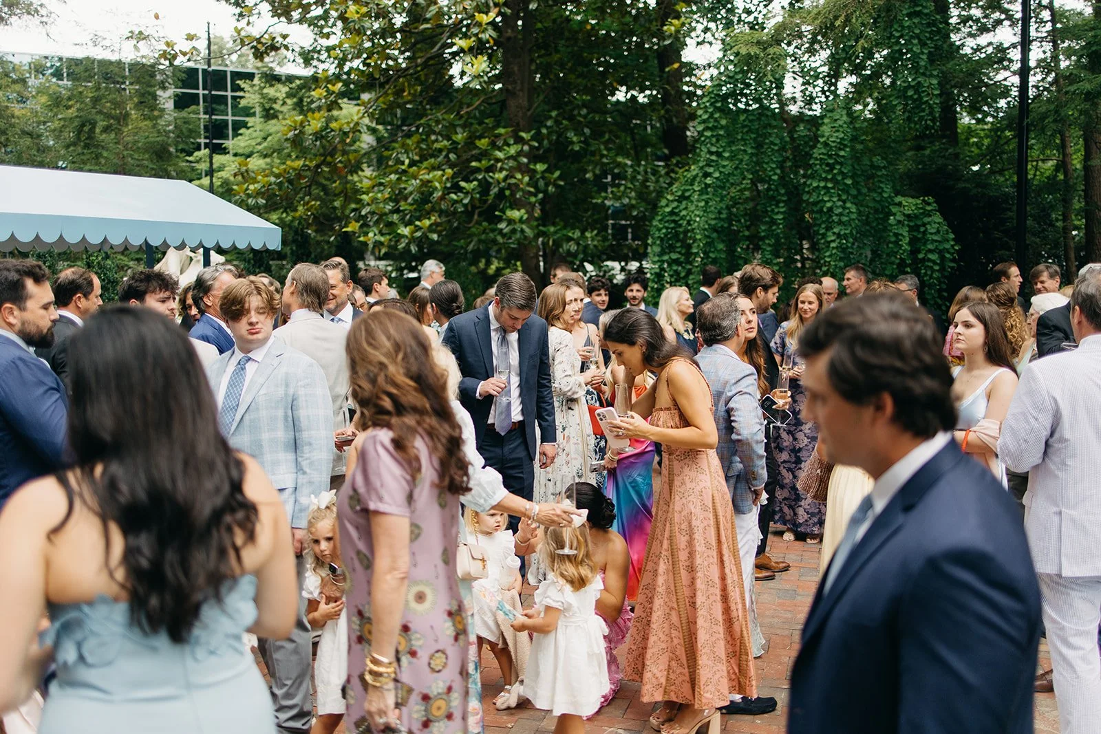 People gathered outdoors at a formal event or celebration, dressed in suits and dresses, with some holding glasses of champagne, surrounded by lush green trees.