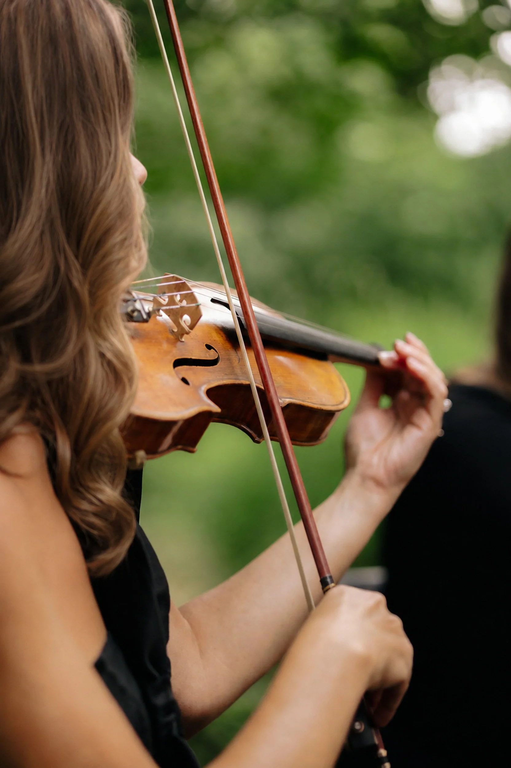 A woman with long, wavy hair wearing a black sleeveless top playing a violin outdoors with a green, blurred background.