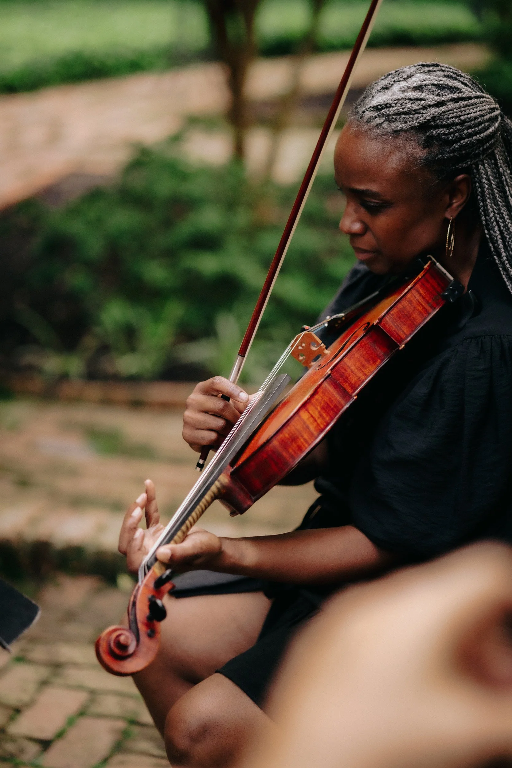 A woman with braided hair playing a violin outdoors, with a blurred green background.