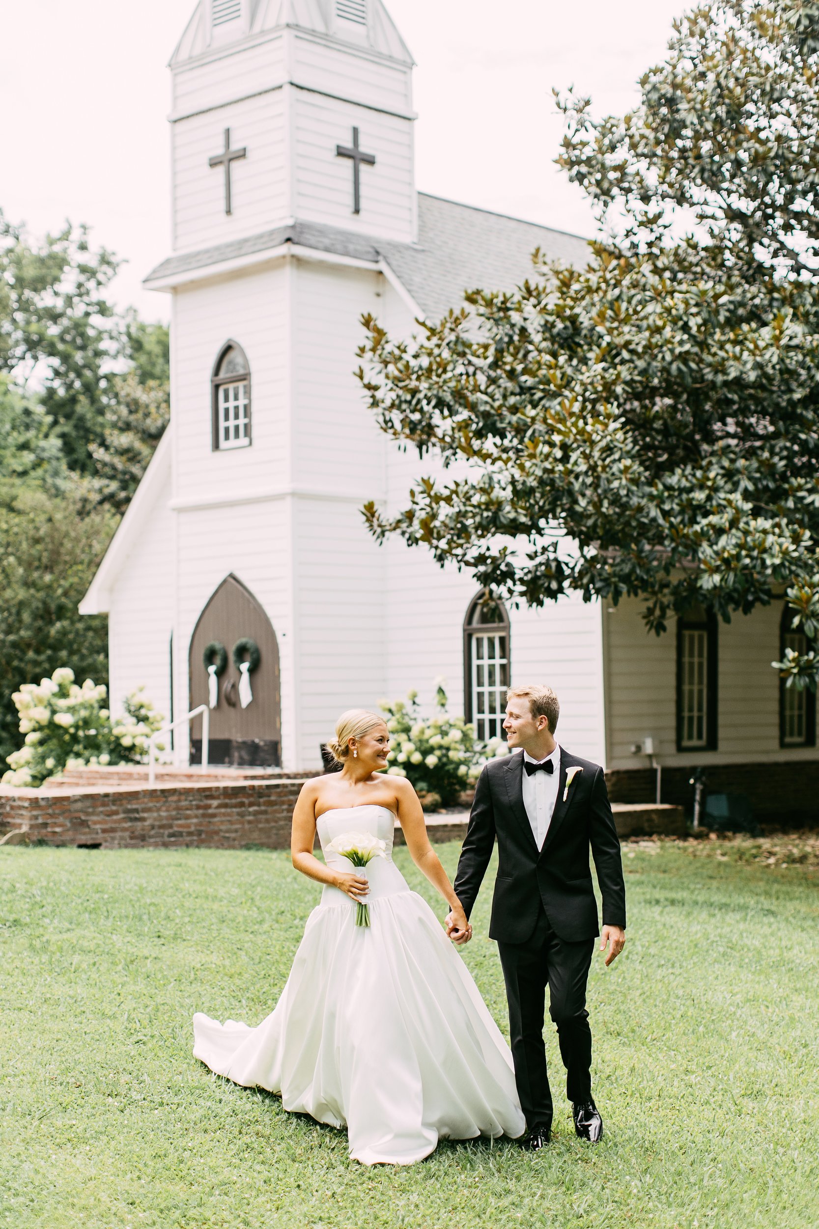A bride and groom smiling and walking hand in hand outside a white church. The bride holds a bouquet of flowers, wearing a strapless wedding dress. The groom wears a black tuxedo. There is greenery and trees around, with a brick pathway and church in the background.