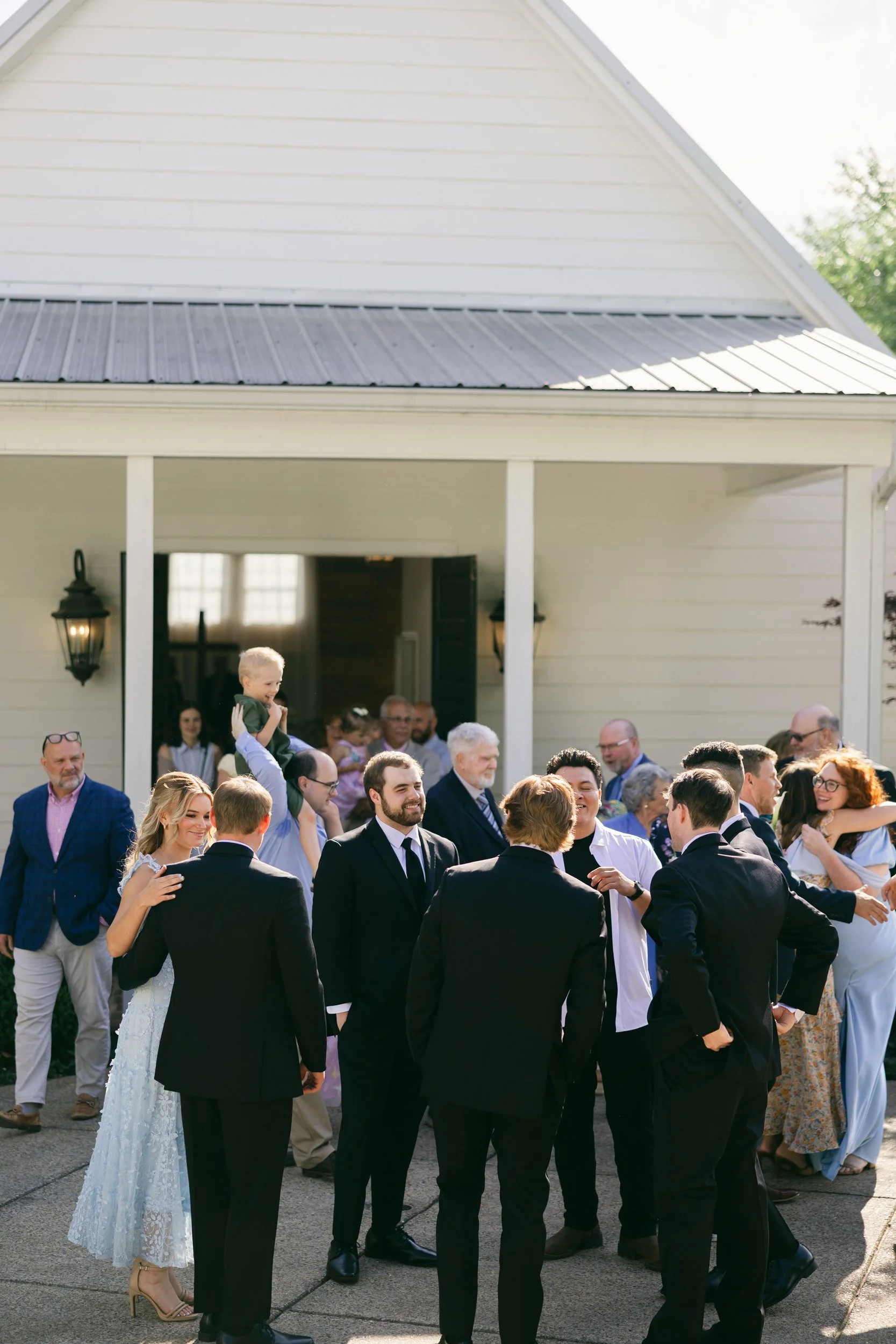 A group of people at an outdoor wedding celebration in front of a white house with a gray roof, with some people dressed in formal attire and a woman in a white lace dress with a bouquet, enjoying the event.