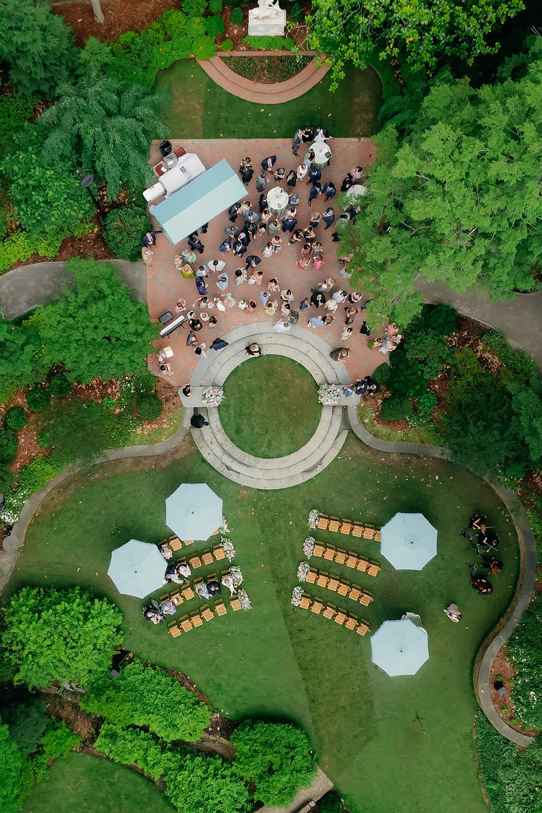 An aerial view of a wedding ceremony in a garden with guests gathered around a white arch, tables, and chairs under umbrellas, surrounded by trees and green foliage.