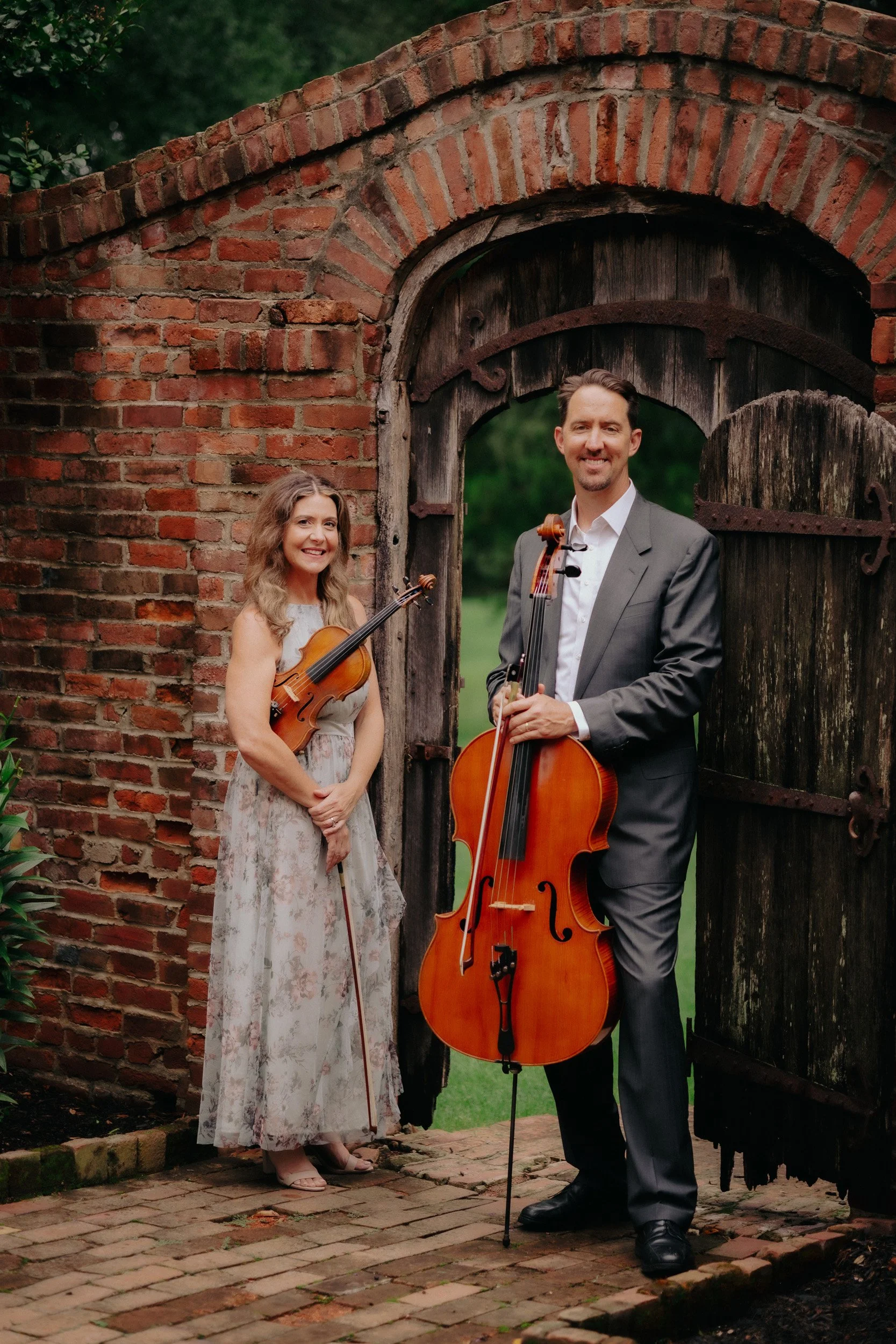 A man in a gray suit and a woman in a floral dress are standing outdoors on a brick pathway in front of a wooden gate and brick wall, holding violins and a cello.