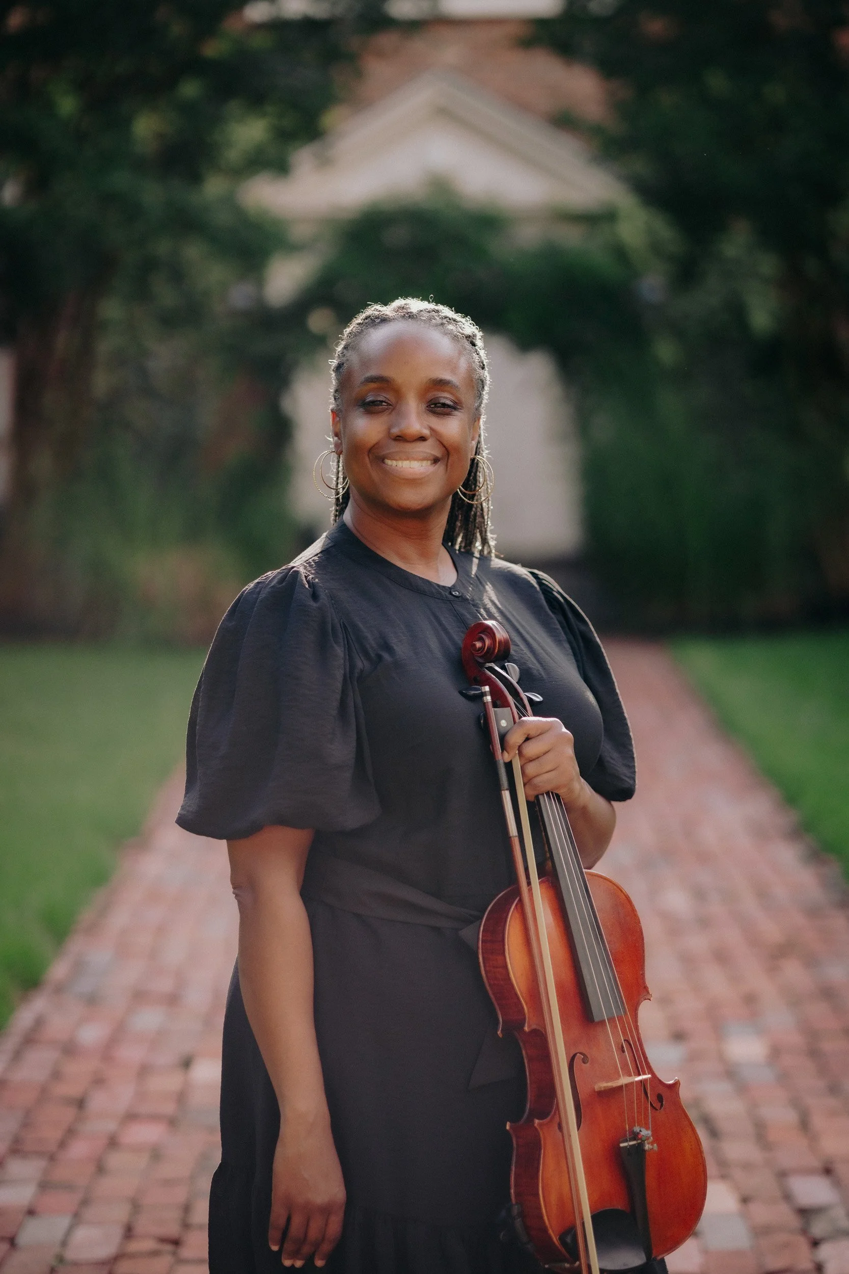 A woman in a black dress smiling, holding a violin, outside on a brick pathway with greenery and trees in the background.