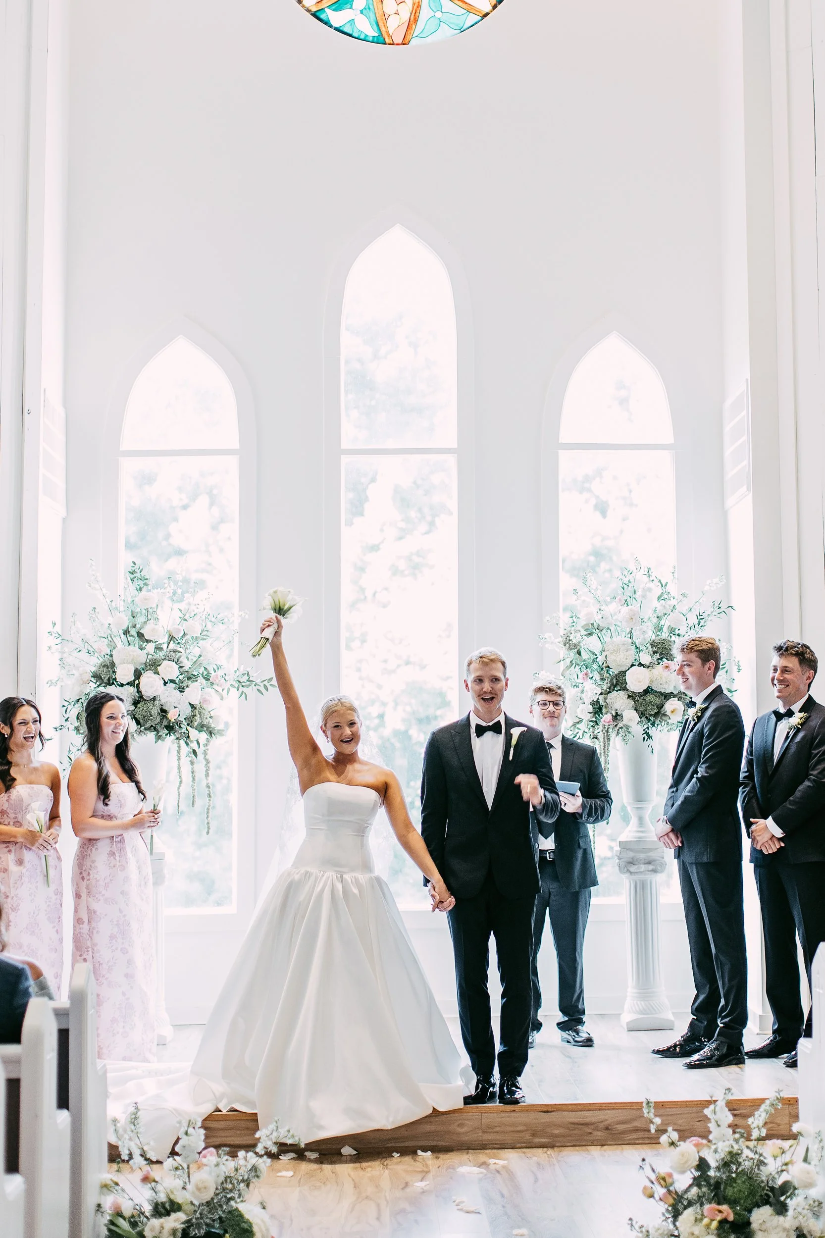 A wedding ceremony taking place indoors with a bride and groom standing hand in hand at an altar. The bride is holding a white bouquet, and the groom is dressed in a tuxedo. Bridesmaids and groomsmen are standing on either side, smiling. The background features large, arched windows and floral arrangements.