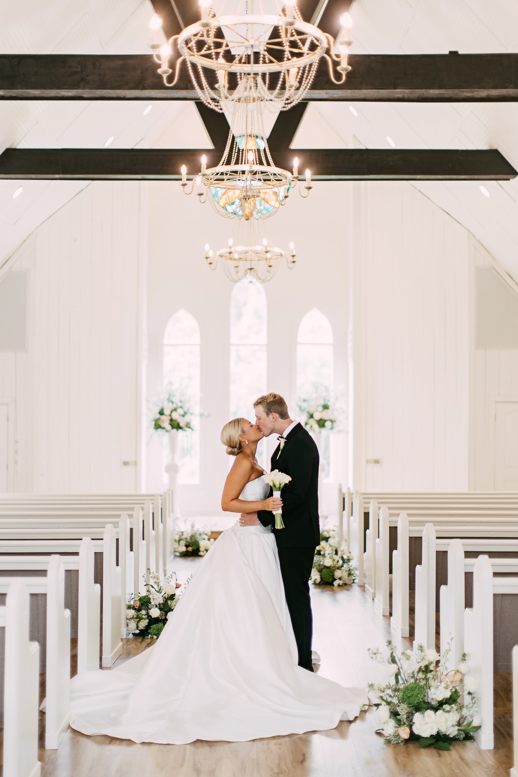 A bride and groom sharing a kiss inside a decorated wedding venue, with the bride in a white gown holding a bouquet, and the groom in a black tuxedo, standing in an aisle lined with white pews and floral arrangements.