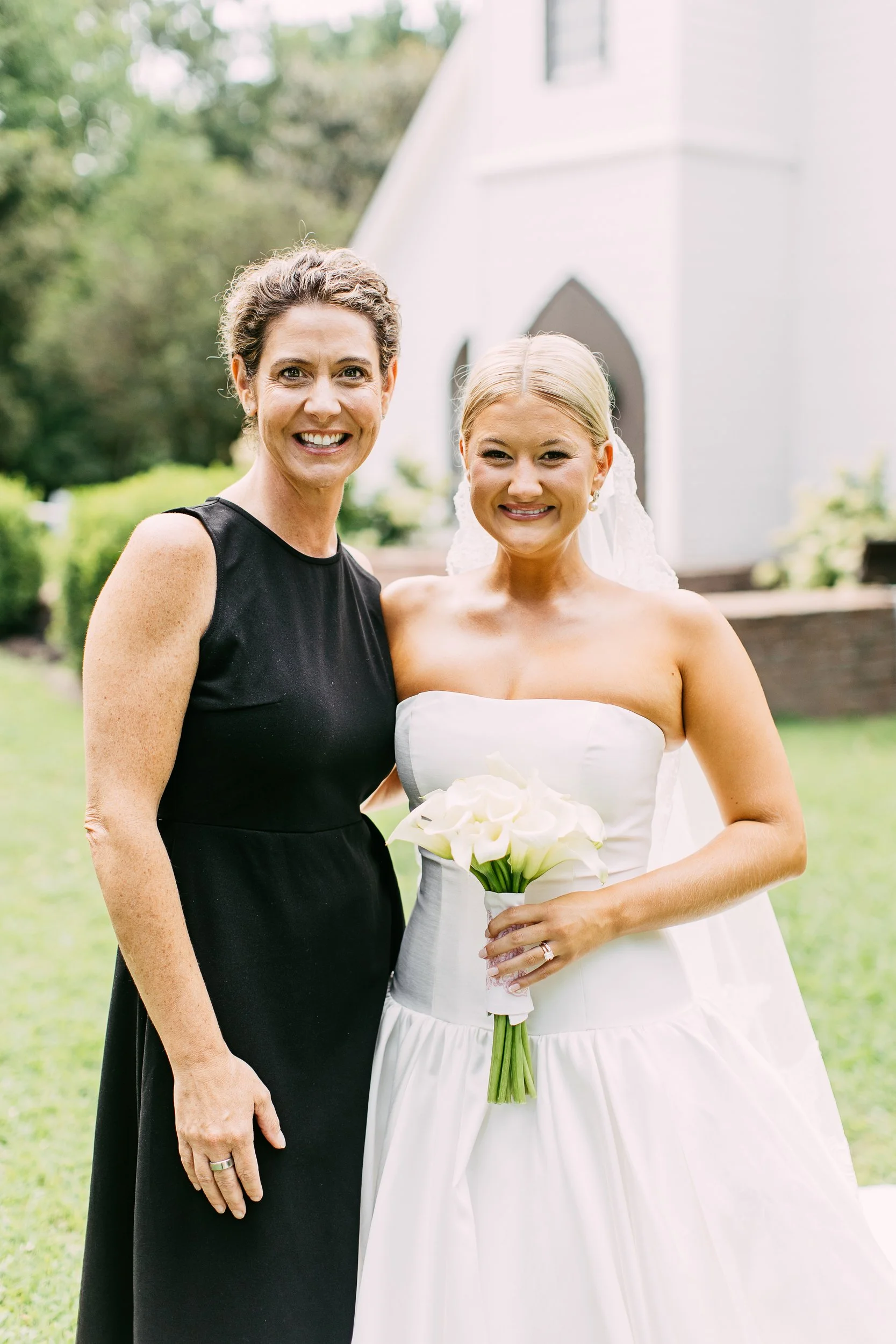 A bride in a white wedding dress holding a bouquet of white calla lilies standing next to a woman in a black dress, outdoors with greenery and a white building in the background.