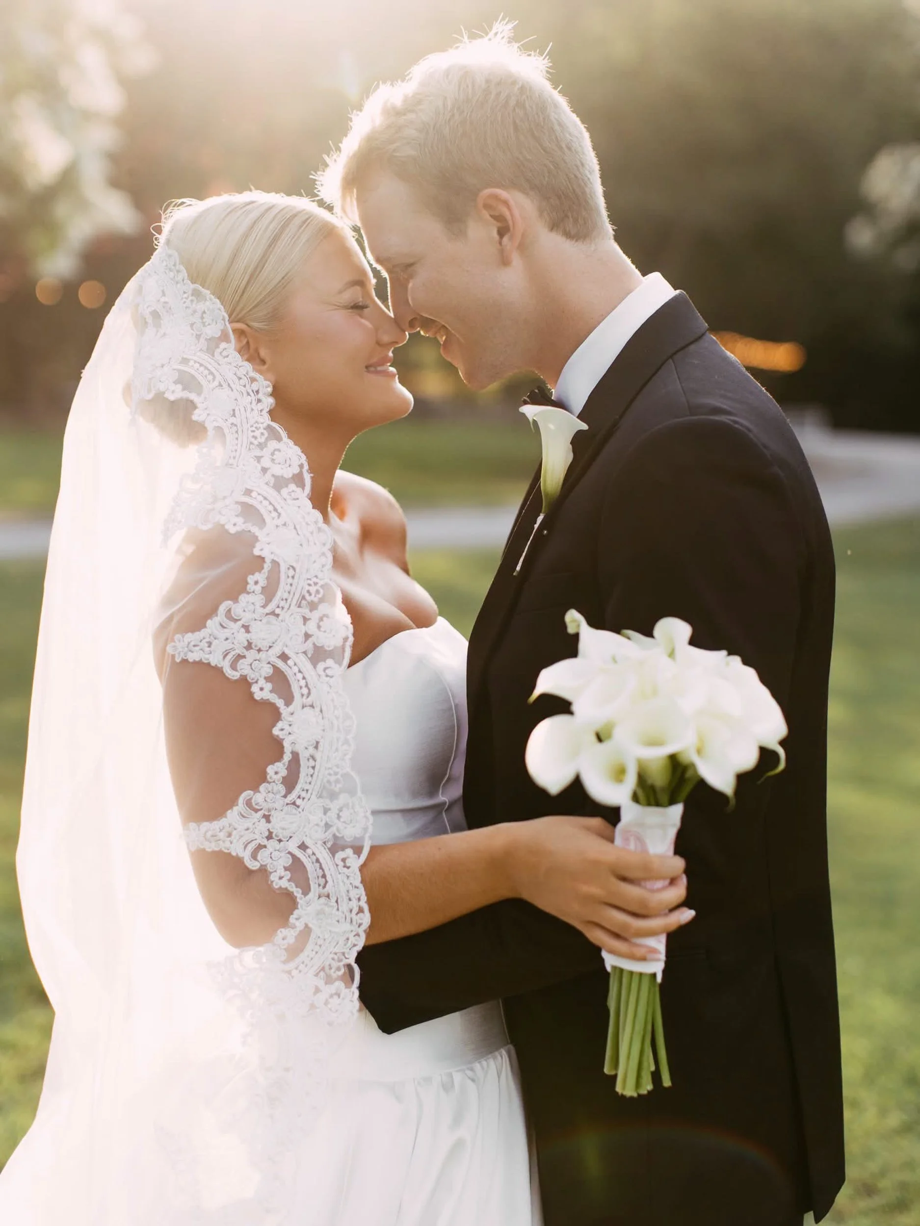 A bride and groom in wedding attire touching foreheads and smiling, outdoors during sunset, with the bride holding a bouquet of white lilies.