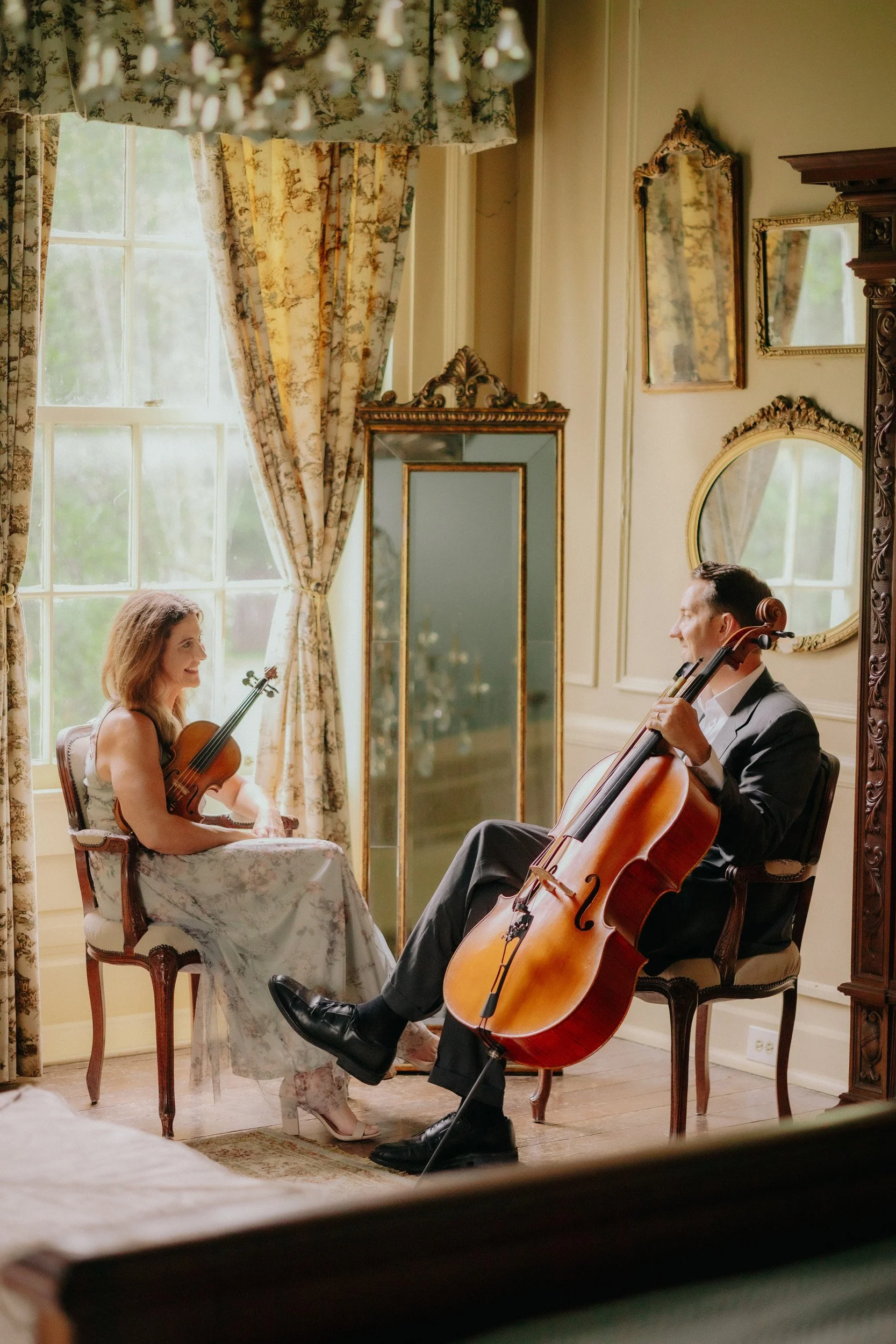 A woman with curly hair playing the violin and a man in a suit playing the cello in a vintage decorated room with large windows, floral curtains, and ornate mirrors.