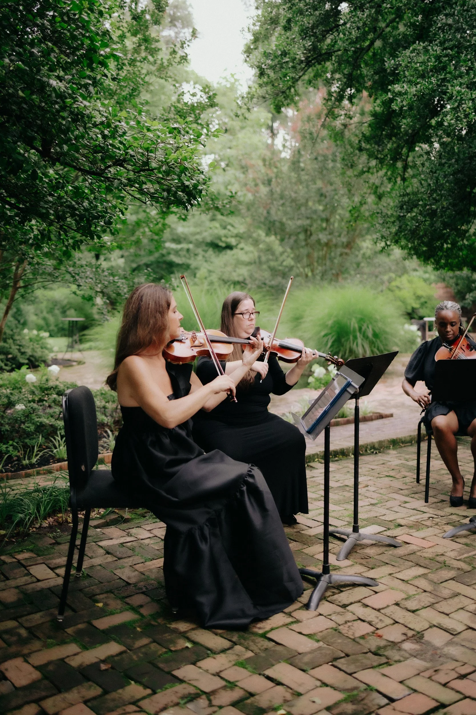Three women are playing violins outdoors in a lush, green garden with trees and brick pathway.