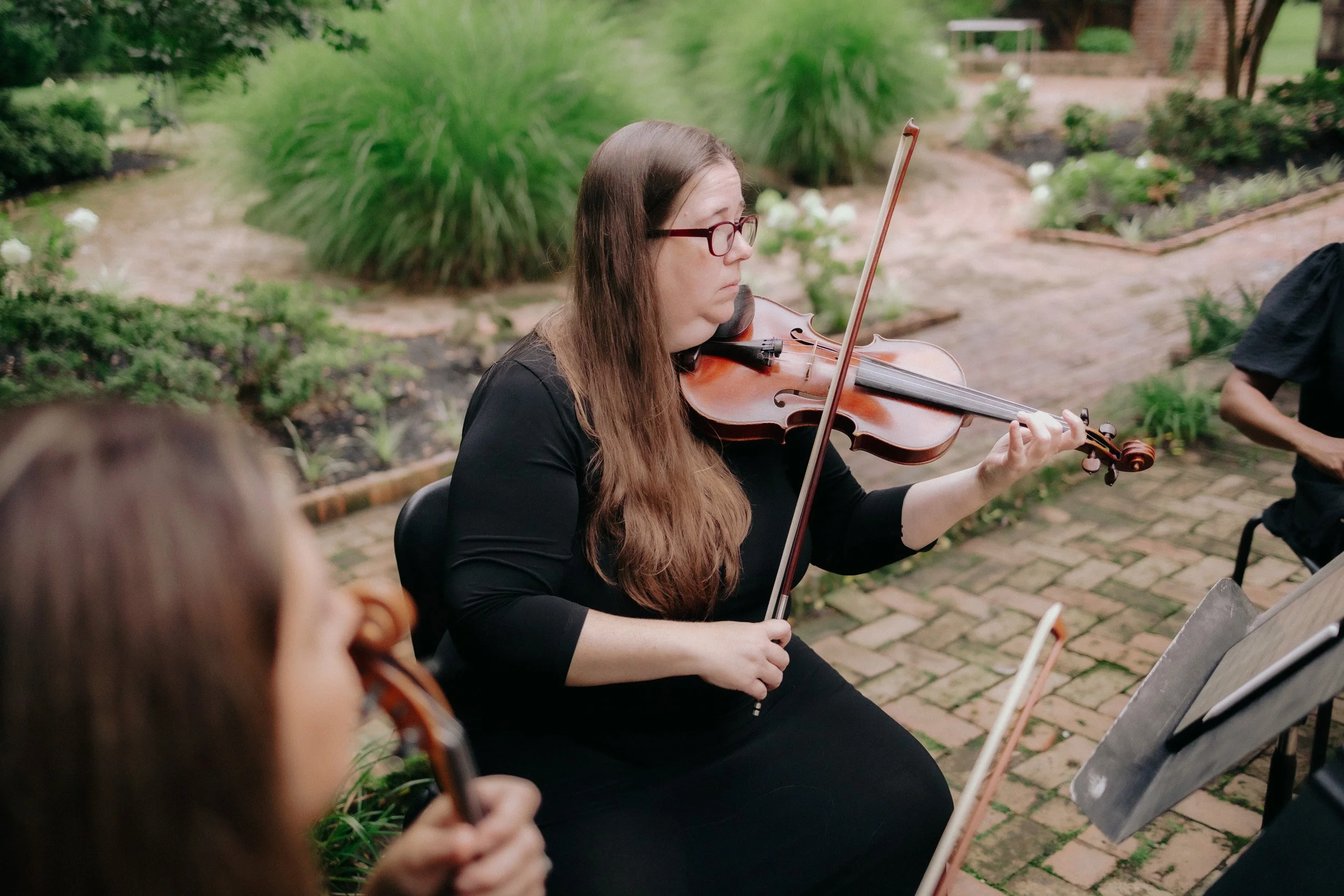 A woman playing the violin outdoors in a garden setting, with other children also playing violins nearby.