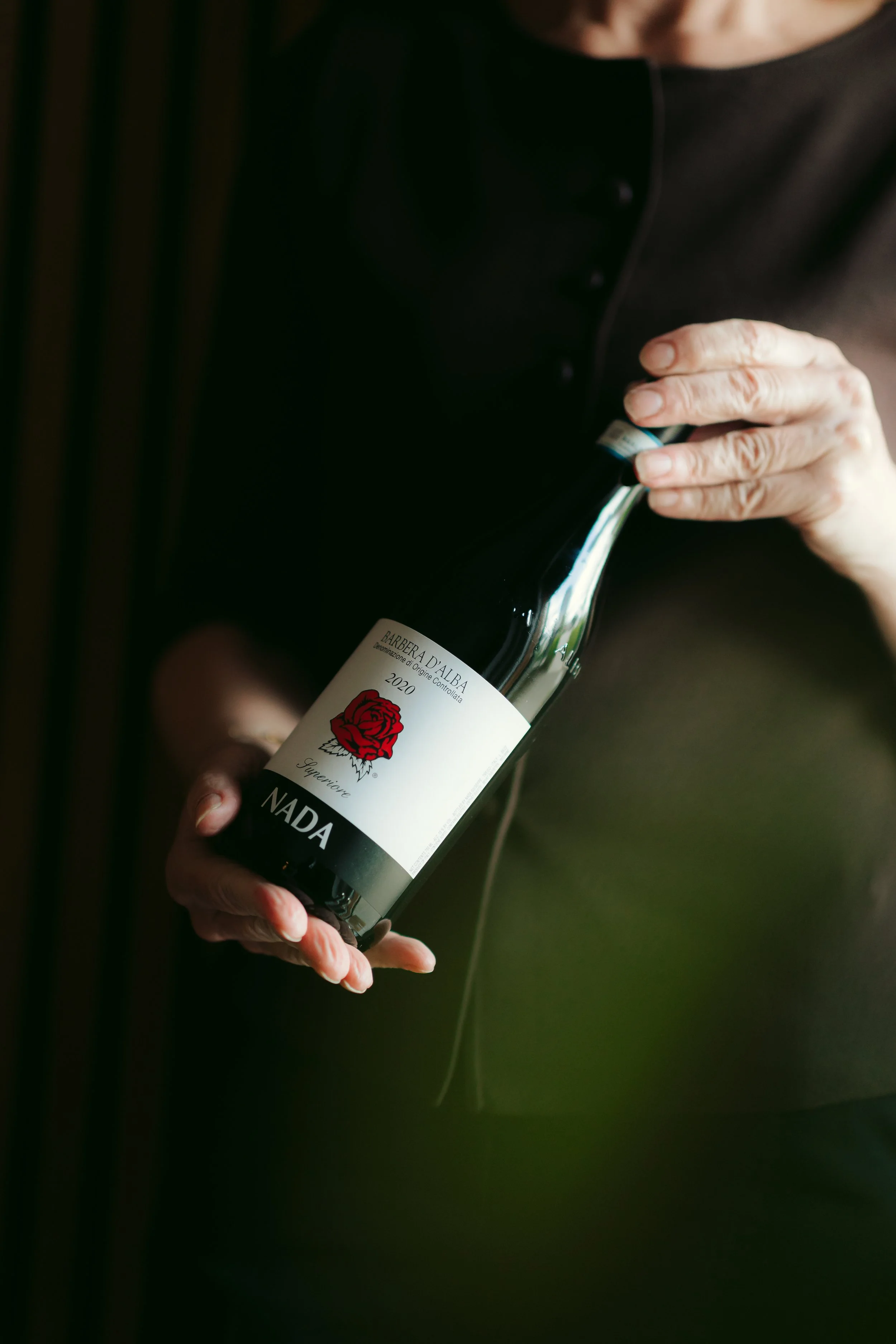 A person's hands holding a bottle of red wine with a white label that has a red rose illustration, and a black outfit visible.