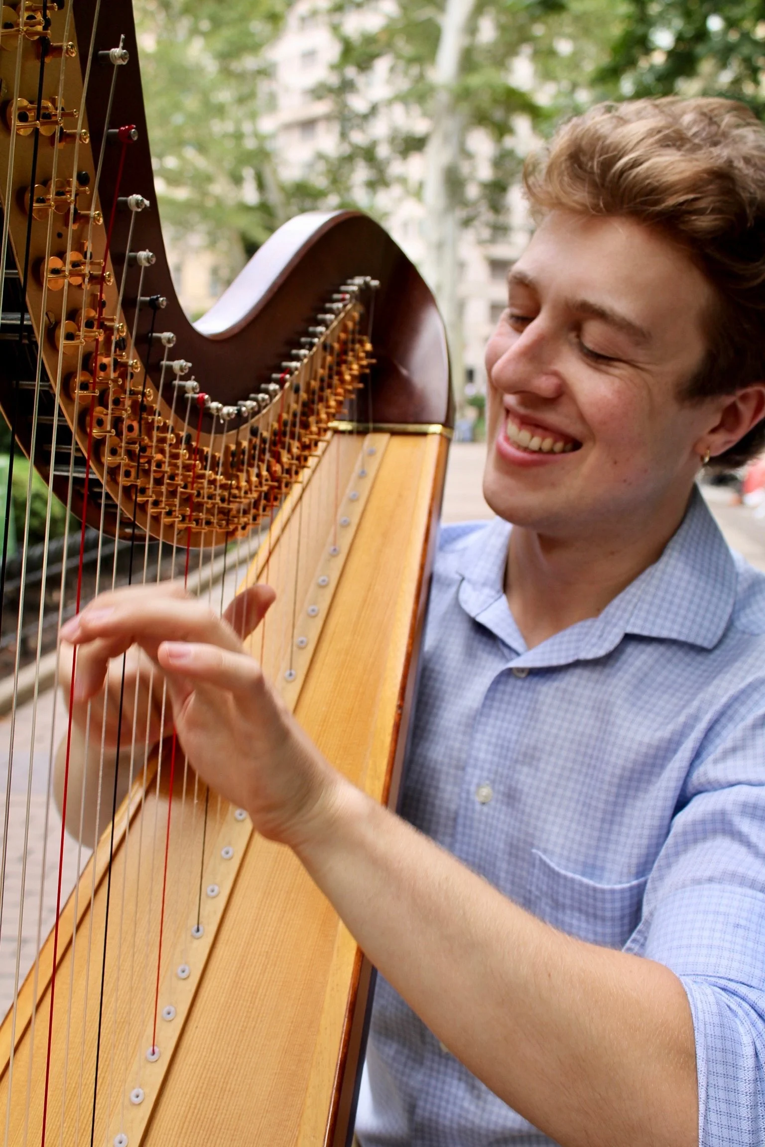 Daniel Benedict playing a harp outdoors, smiling with closed eyes, in a park with trees and buildings in the background.