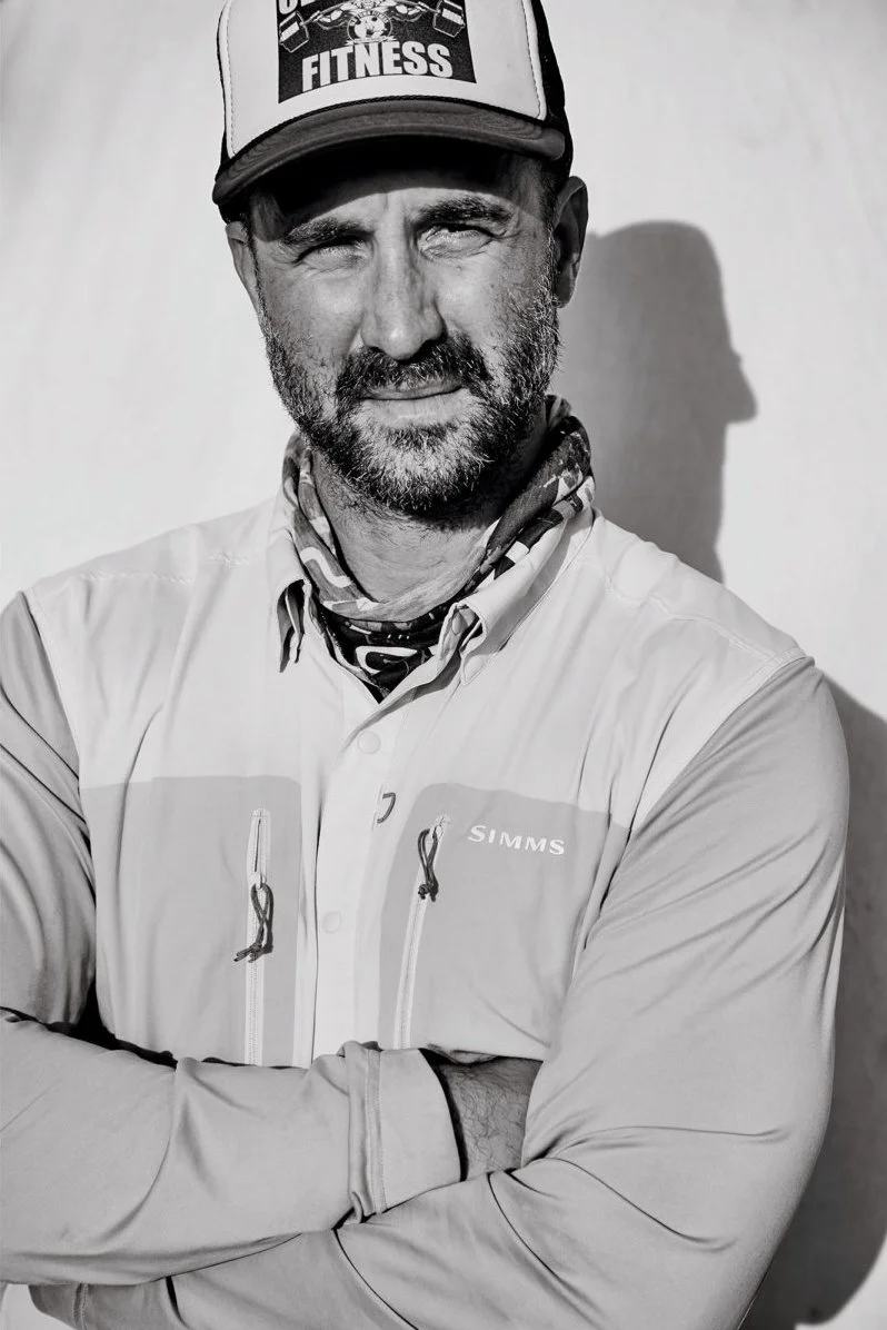 Black and white photo of a Nathaniel wearing a cap with 'FITNESS' written on it, a scarf around his neck, and a jacket with the brand name 'SIMMS.' He is standing with arms crossed in front of a plain background.