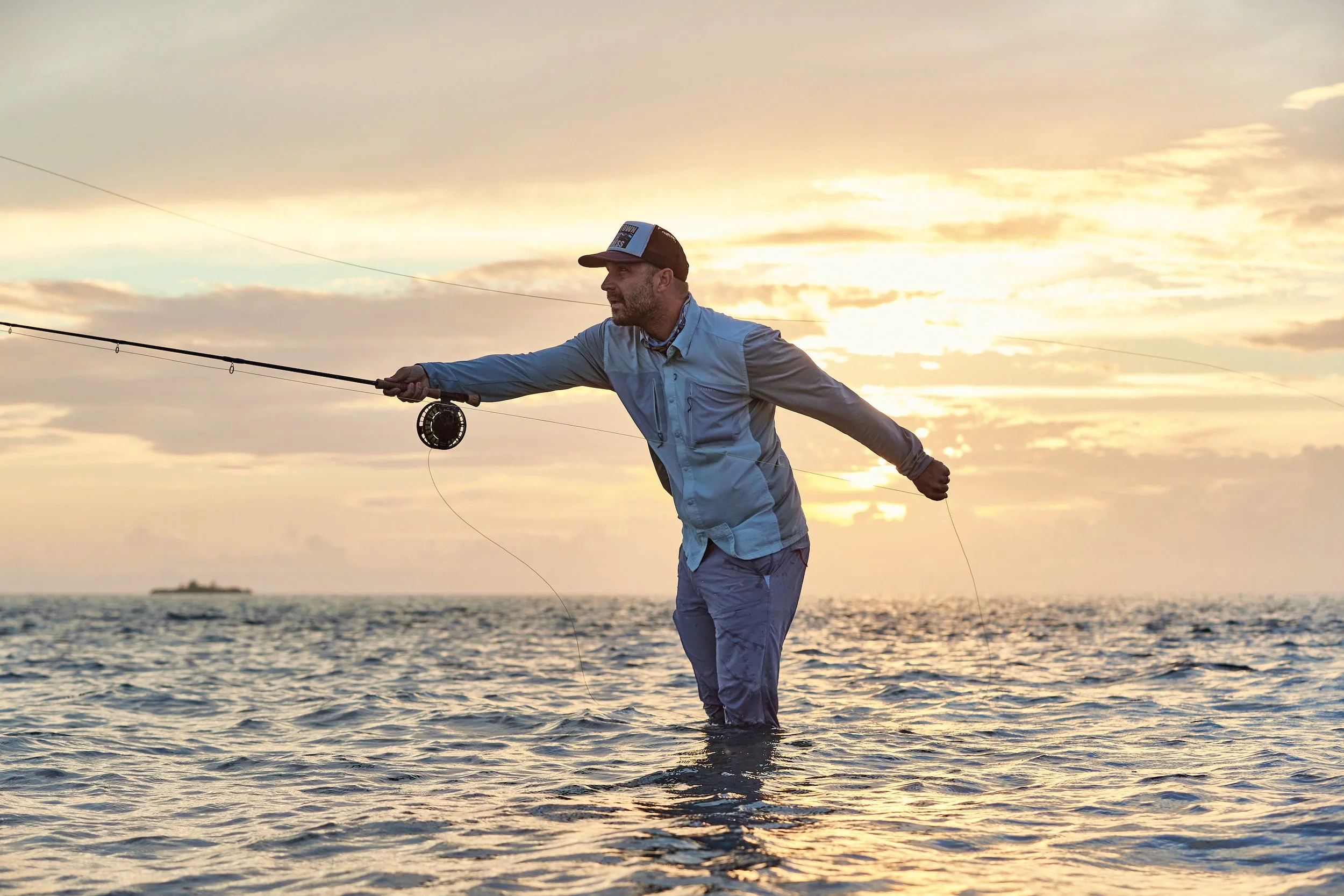 Nathaniel fly fishing in the water at sunset or sunrise, wearing a cap and a light jacket, with a fishing reel in his hand. There is a ship or boat visible in the background on the horizon.
