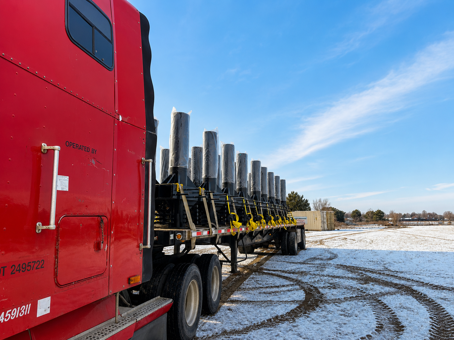 A large red semi-truck with wind turbine blades loaded on the flatbed trailer, parked in a snowy open field under a blue sky.
