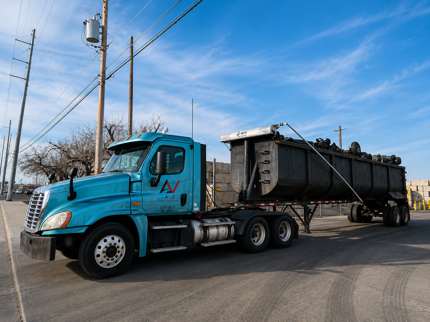 Blue semi-truck with a black dumpster trailer loaded with debris parked on a street under a partly cloudy sky.
