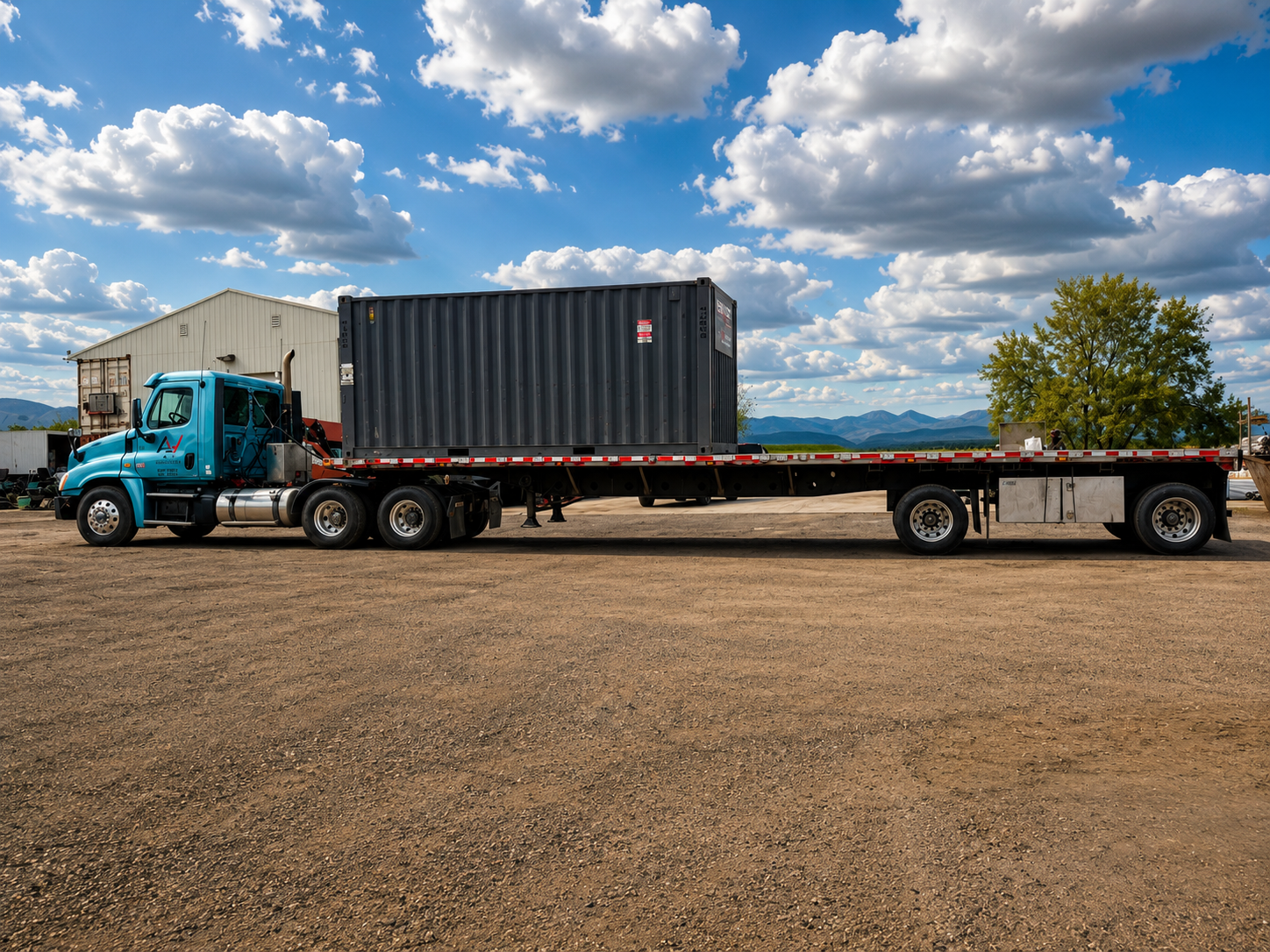 Blue semi-truck with a black shipping container on a flatbed trailer parked on a dirt lot, with a tree, a white building, and mountains in the background under a partly cloudy sky.