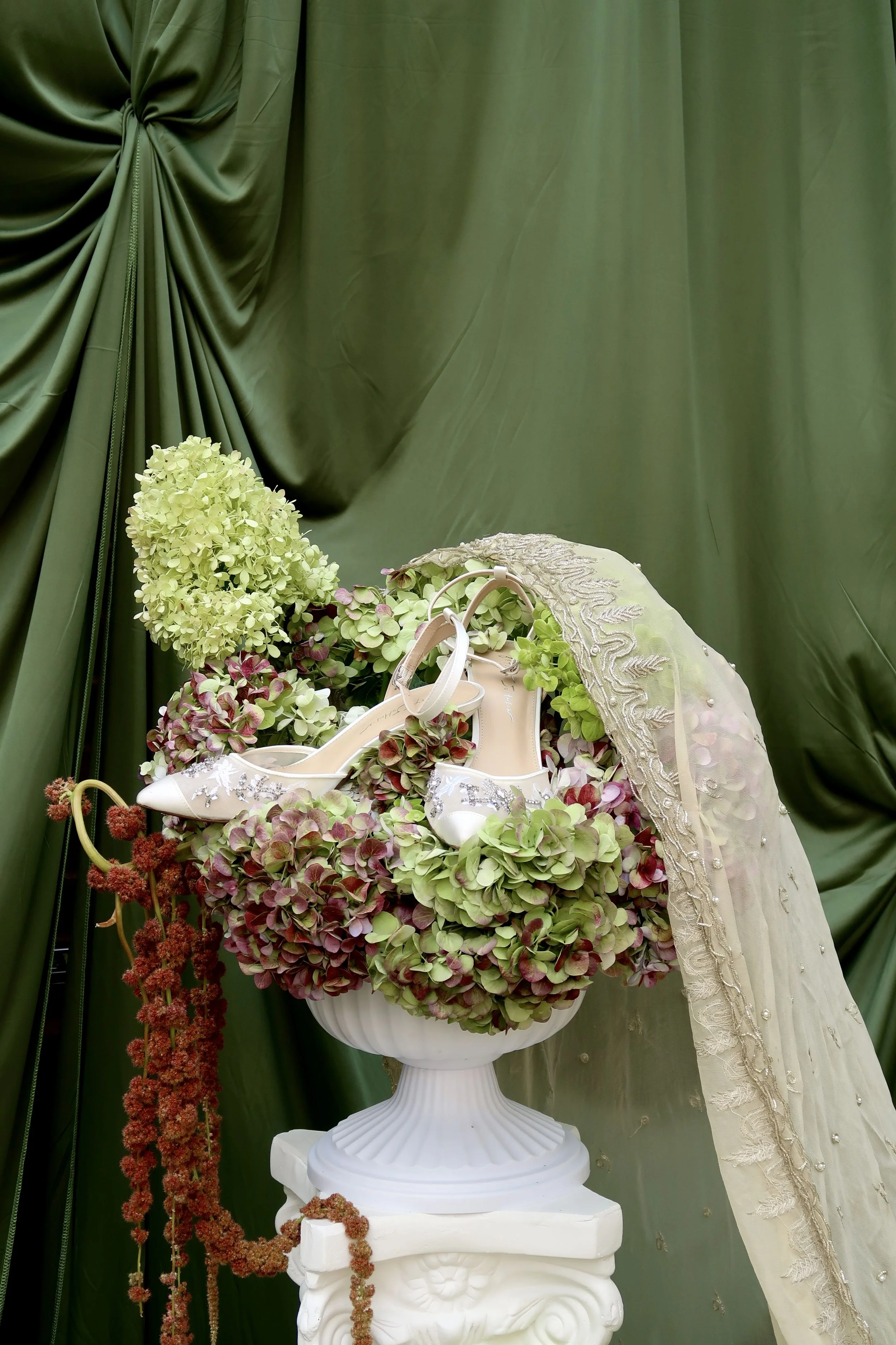 A pair of white high-heeled shoes with floral embellishments displayed in a white vase surrounded by green and pink hydrangea flowers, set against a green satin curtain backdrop with a sheer lace fabric draped on the side.