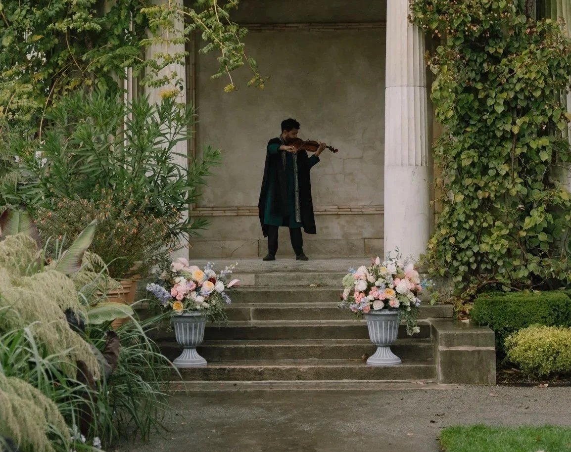 A man playing the violin on a small outdoor stage with flowers in vases on the steps, surrounded by greenery and columns.