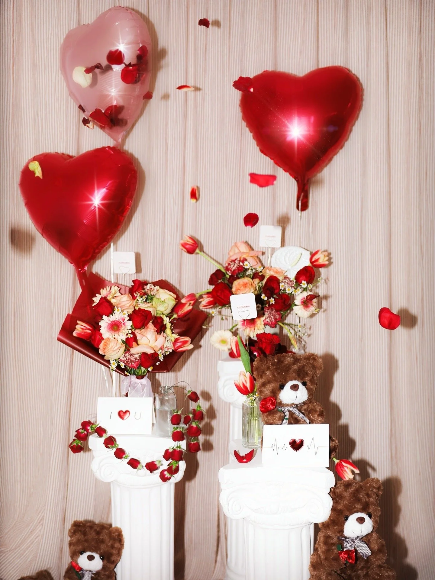 A Valentine's Day themed arrangement with three white classical-style pillars holding vases of mixed flower bouquets, surrounded by red and brown teddy bears, heart-shaped balloons, and scattered rose petals on a beige striped background.