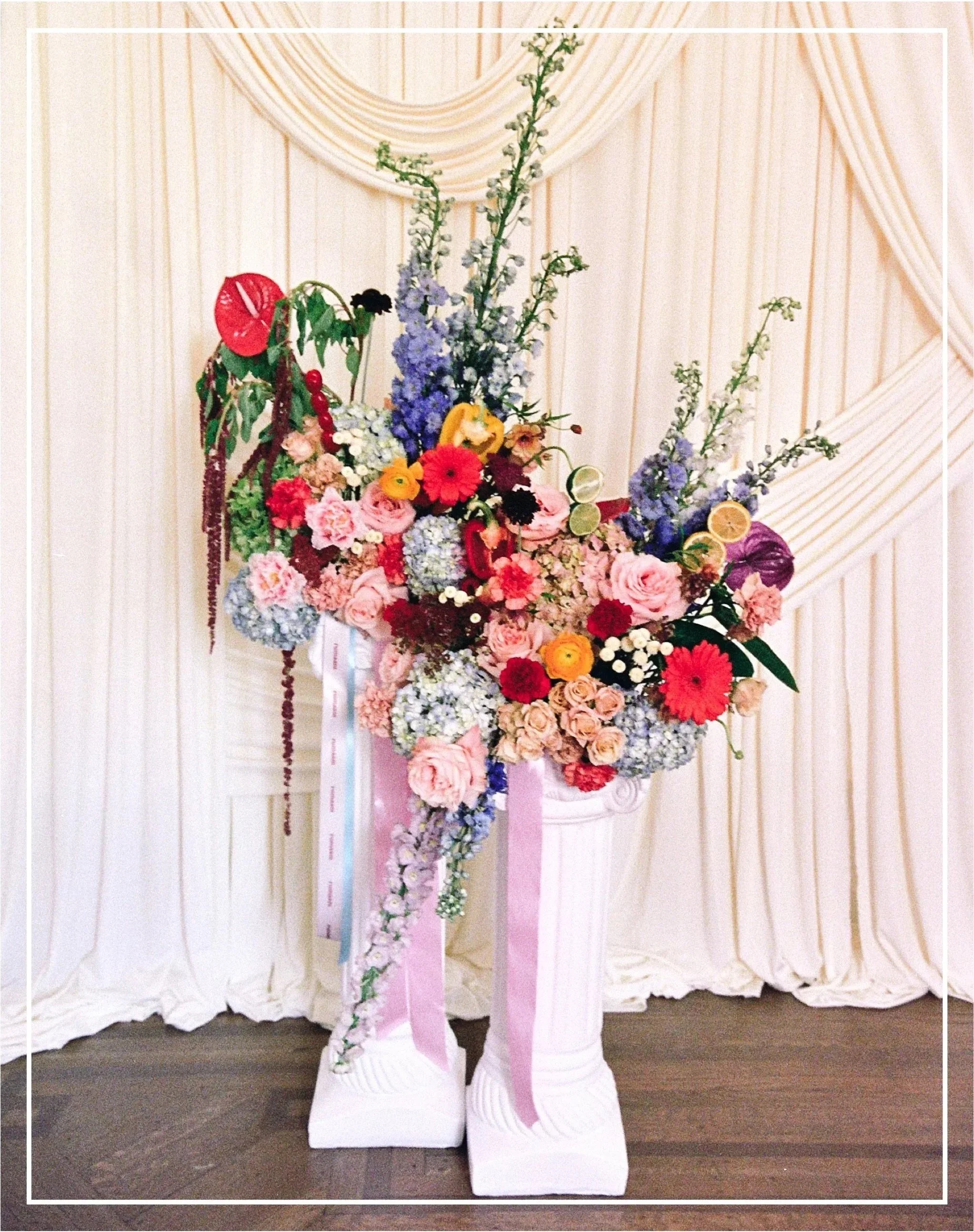 A floral arrangement with various pink, purple, red, yellow, and white flowers, and orange slices, placed on white pedestal bases with pink and white ribbons against a cream curtain background.