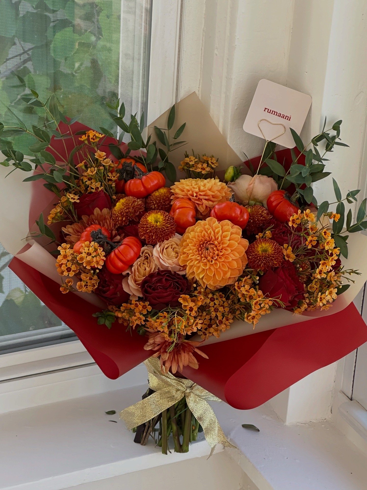 Bouquet of orange dahlias, red roses, small yellow flowers, and green leafy foliage wrapped with red paper and a gold ribbon.