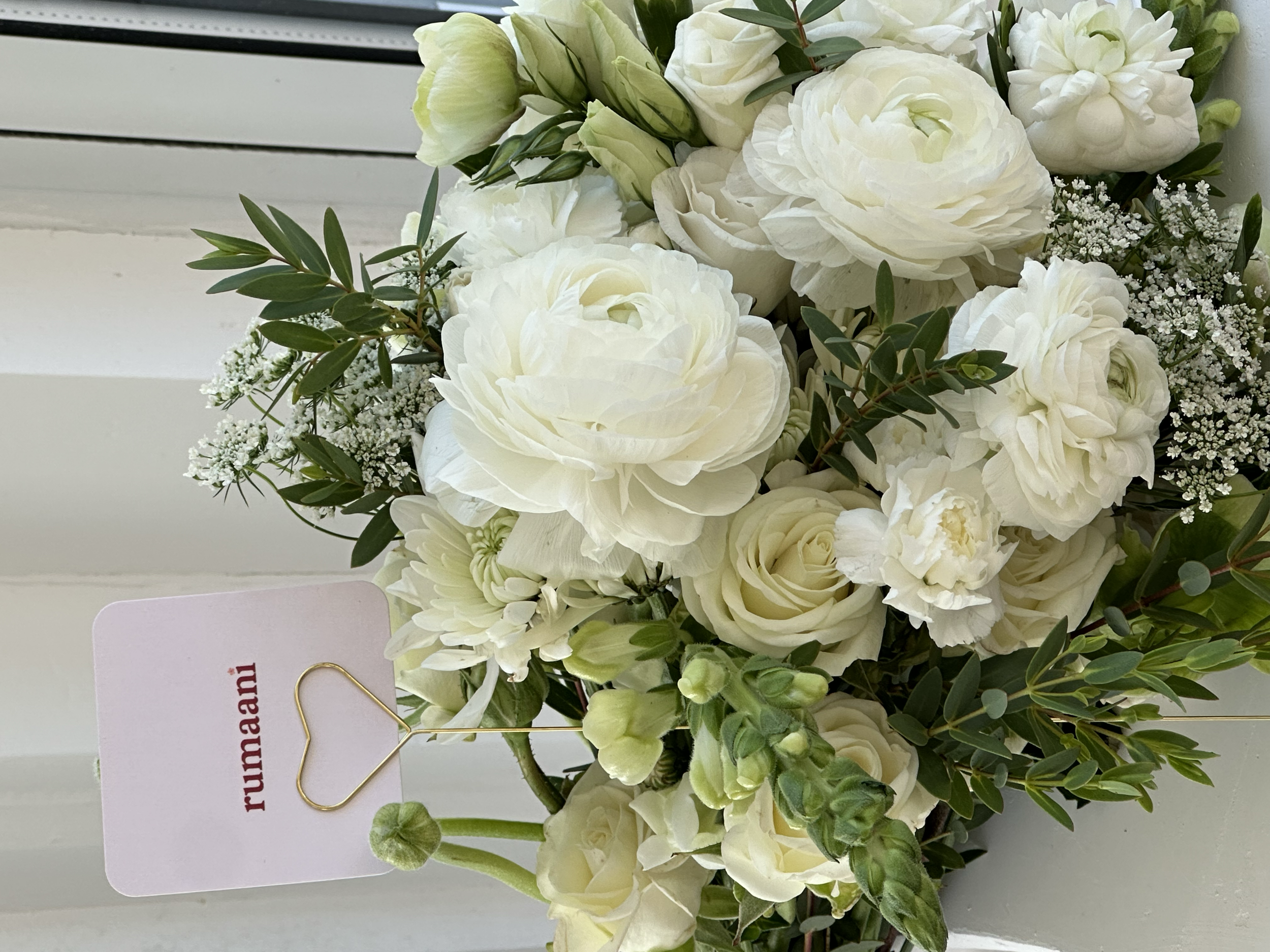 A bouquet of white and cream-colored flowers, including roses, ranunculus, and small filler flowers, with greenery interspersed. The bouquet is placed on a white surface with part of a card visible in the background.