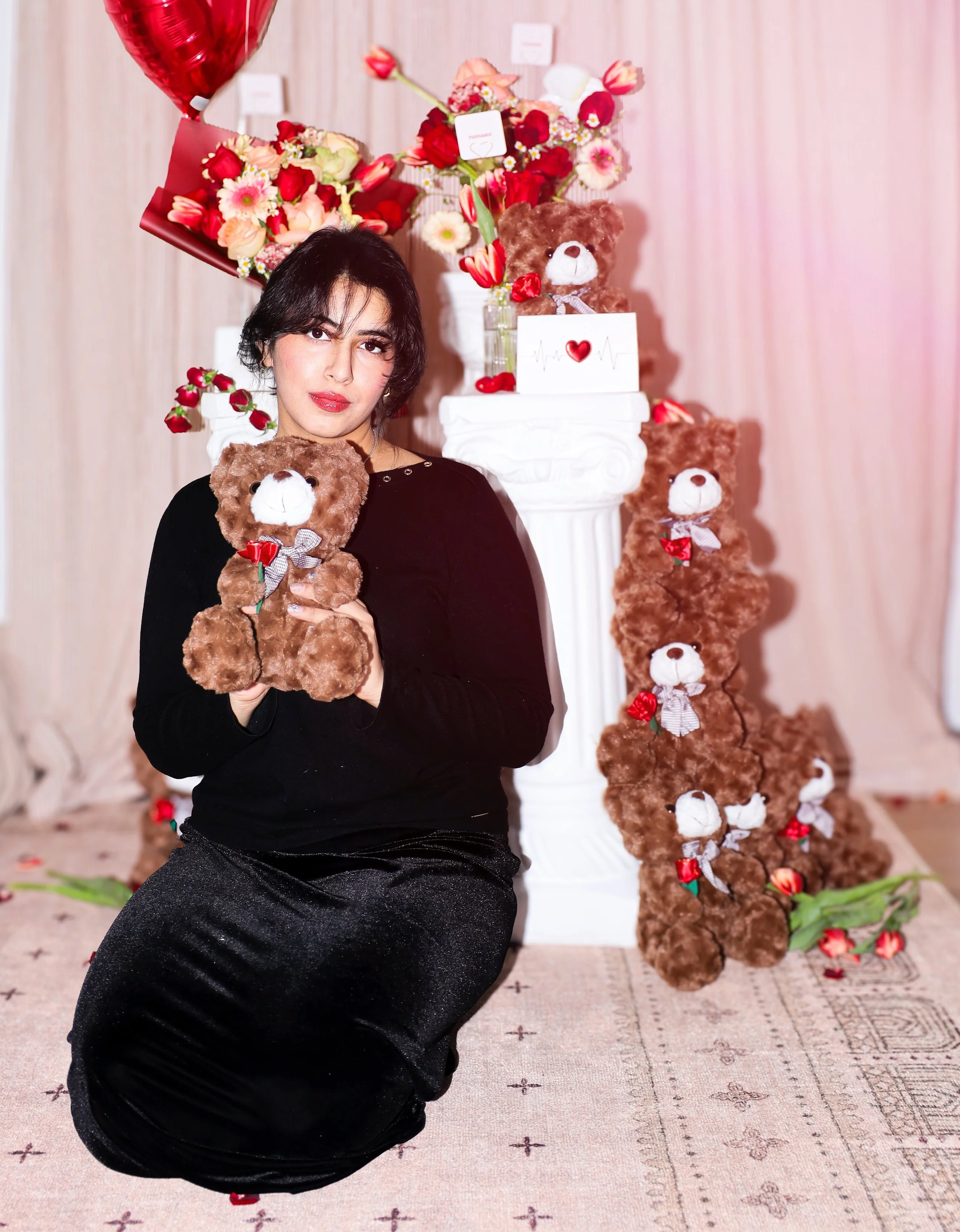 A woman with short black hair sitting on the floor, holding a brown teddy bear with a red flower and gray ribbon. Behind her, there is a white pedestal with a glass vase of colorful flowers and teddy bears with red bows. To the right, a display of several brown teddy bears with bows are stacked vertically, and there are small red flowers scattered around. The background is light pink with wooden paneling.