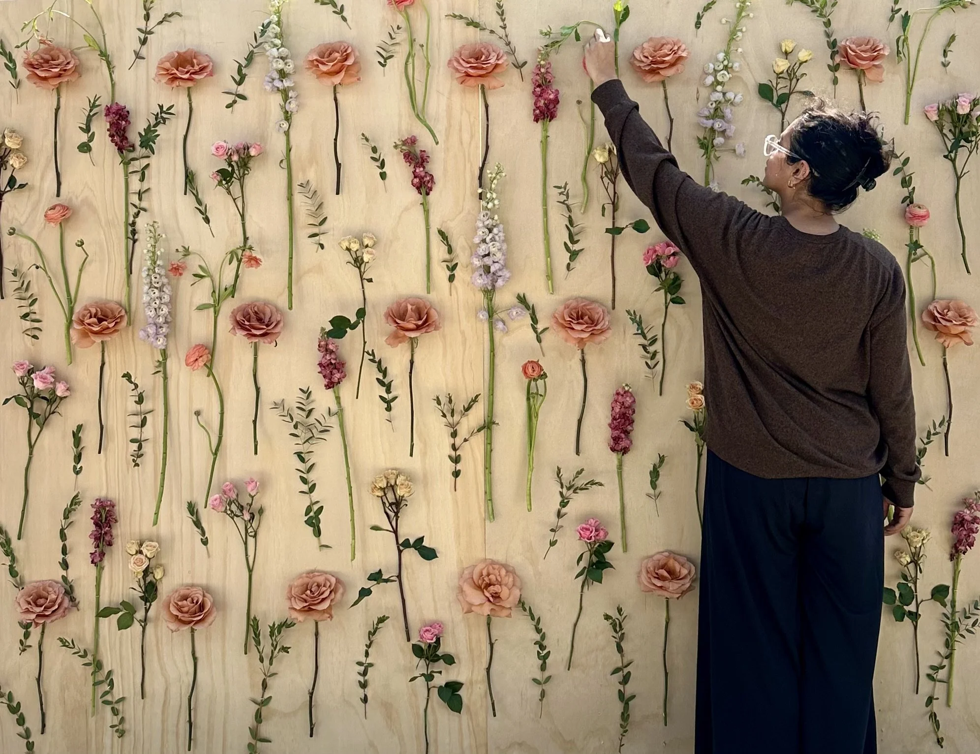 A person with dark hair and glasses arranging flowers on a wall decorated with various types of pink, white, and cream-colored flowers and green foliage.